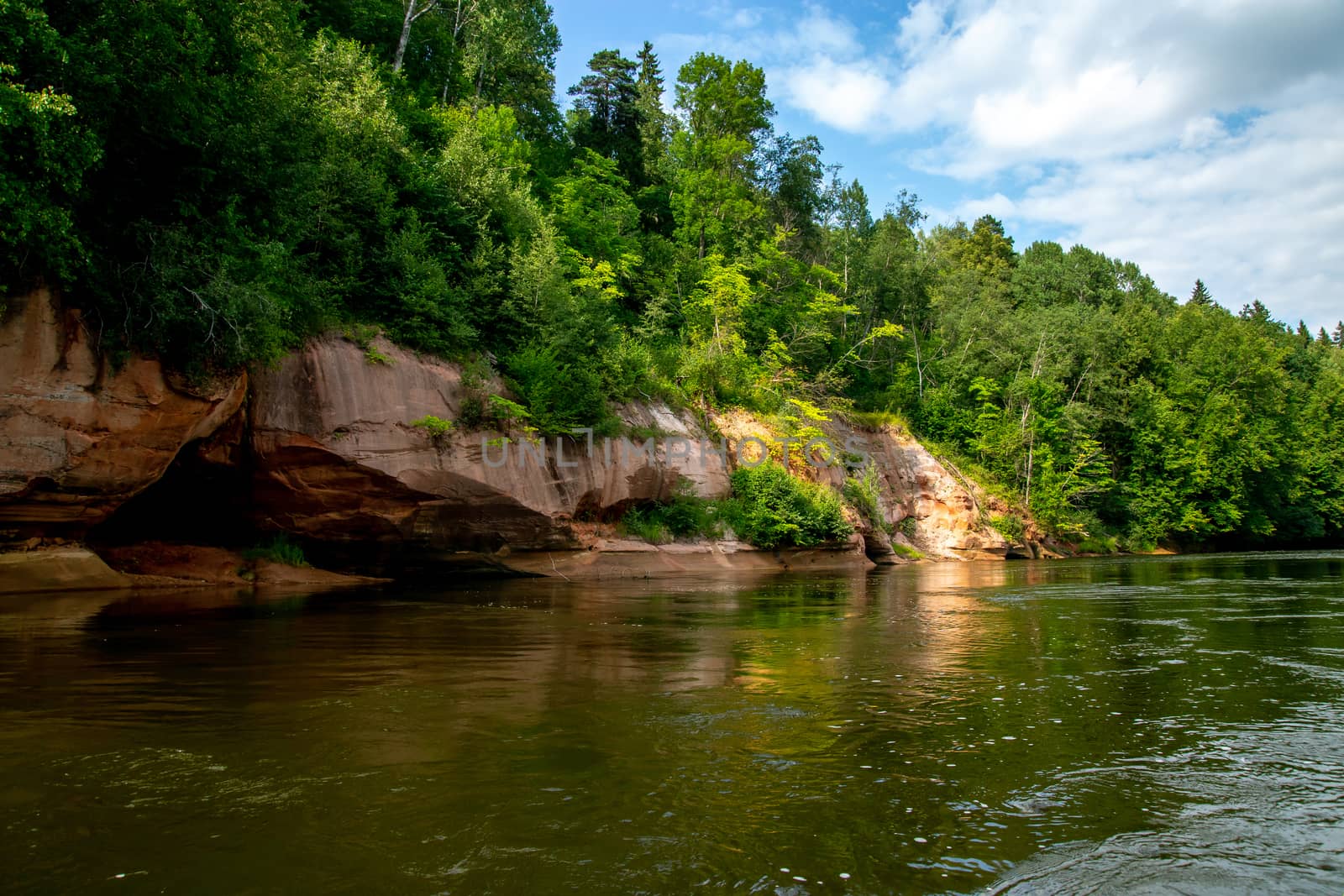 Landscape with cliff near the river Gauja, forest and clouds reflection in water. Cave in cliff. The Gauja is the longest river in Latvia, which is located only in the territory of Latvia. 

