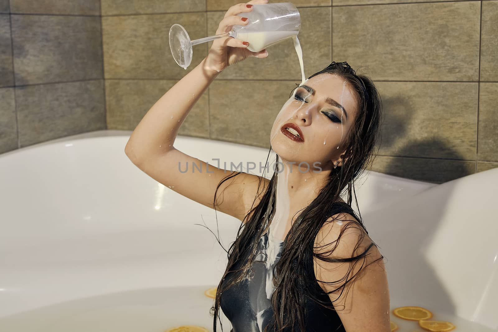 Young woman pours milk on herself. Woman with smeared makeup dressed in the black bathing suit posing in the bath of milk. Conceptual fashion photography for design. Young woman for lifestyle design.