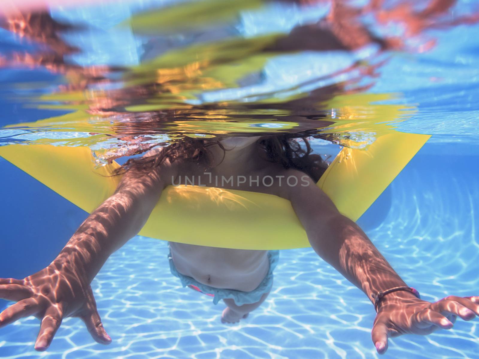 Underwater view of a girl swimming towards the camera with her yellow float in the pool, summer sunlight reflected on the bottom of the pool and there are also reflections on the surface of the water