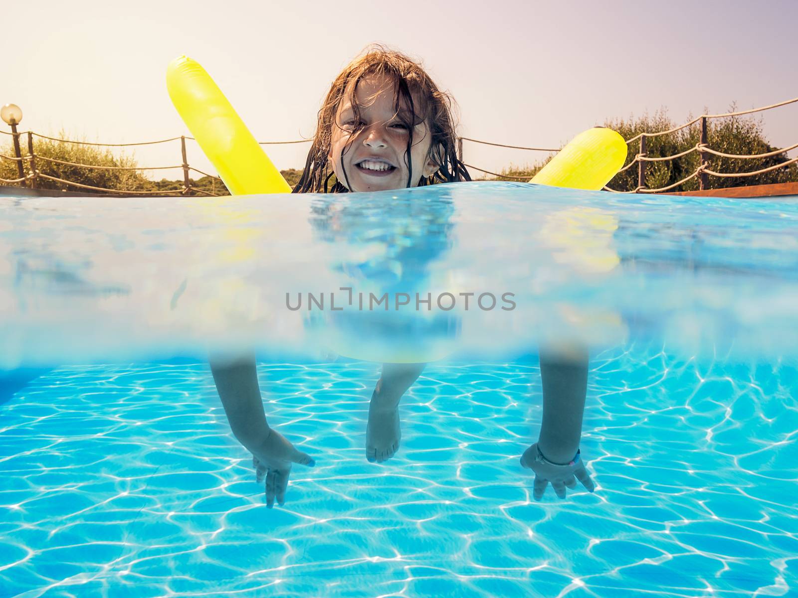 Underwater view of a girl swimming smiling toward the camera with her yellow float in the pool, behind her the glow of the summery sun gleams golden