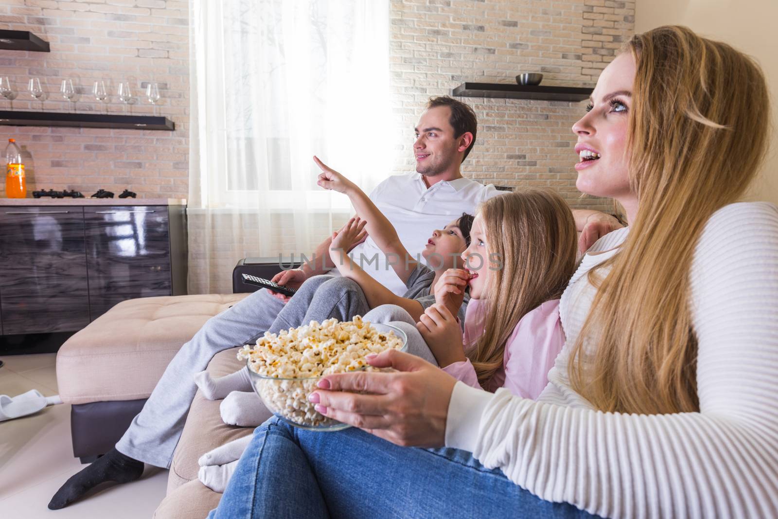 Beautiful young parents and their children are watching TV, eating popcorn and smiling while sitting on couch at home