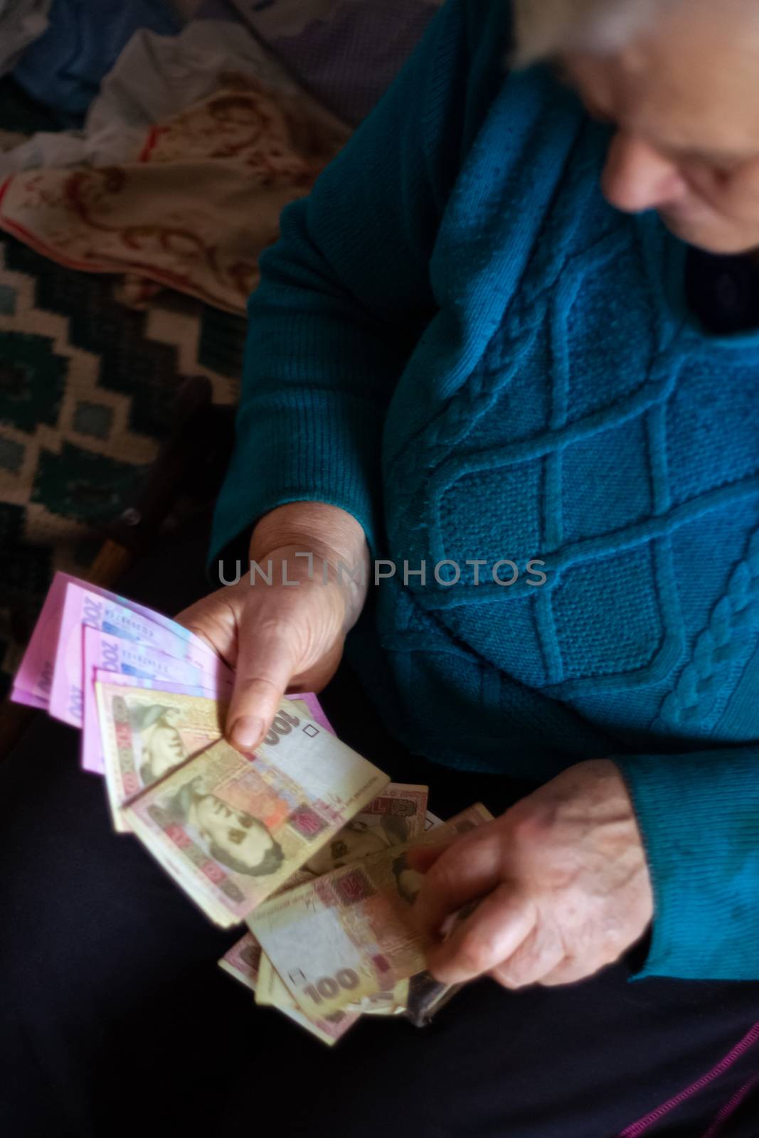 Old poor gray hair woman holds Ukrainian hryvna money in her hands. Woman is sad. Poor life in village. Old age not good. Low-light photo. by alexsdriver