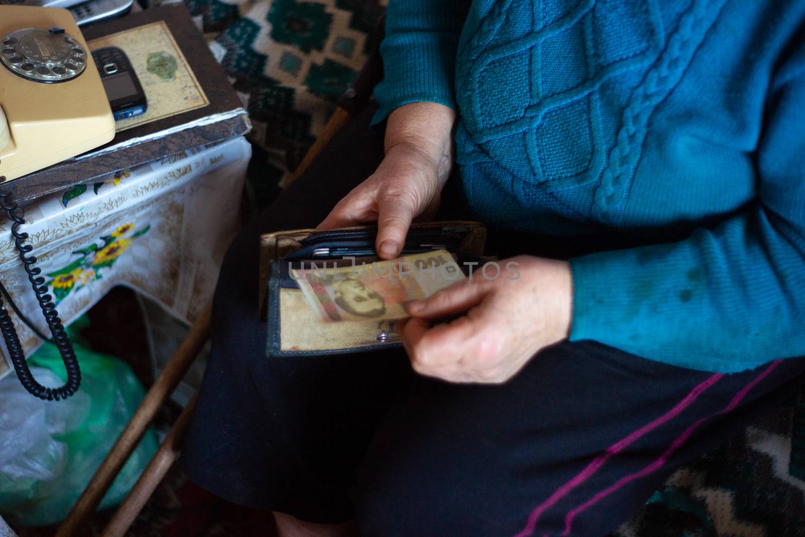 Old poor gray hair woman holds Ukrainian hryvna money in her hands. Woman is sad. Poor life in village. Old age not good. Low-light photo. by alexsdriver
