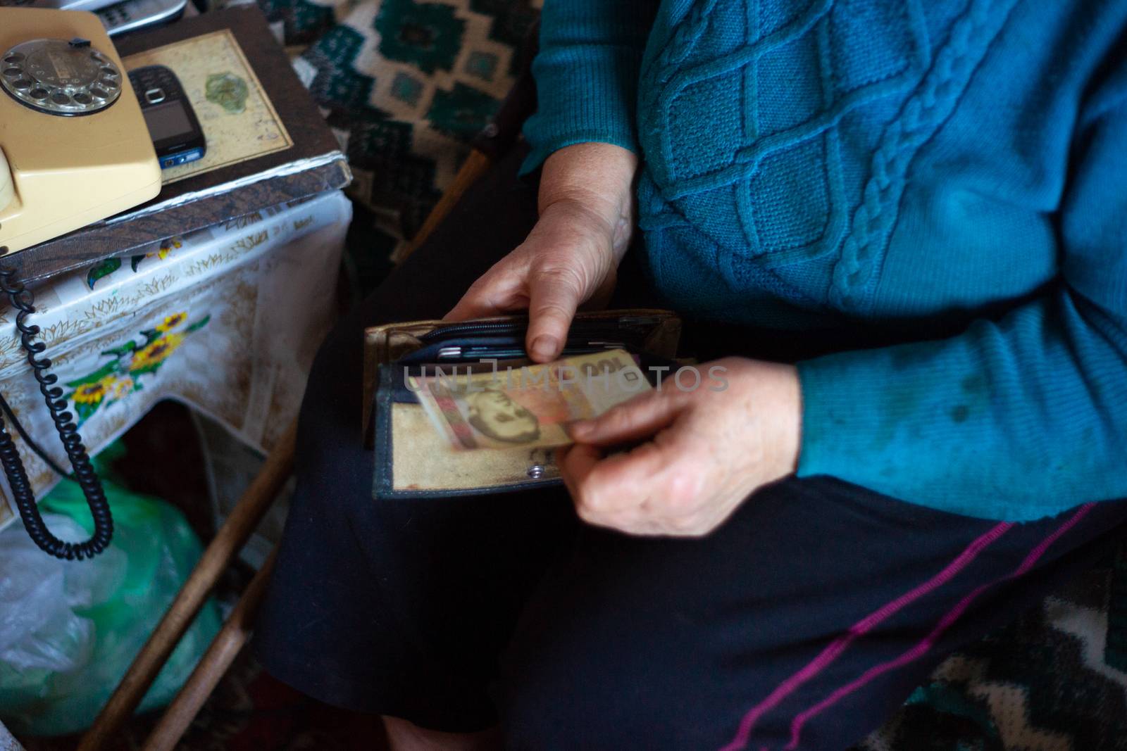 Old poor gray hair woman holds Ukrainian hryvna money in her hands. Woman is sad. Poor life in village. Old age not good. Low-light photo.