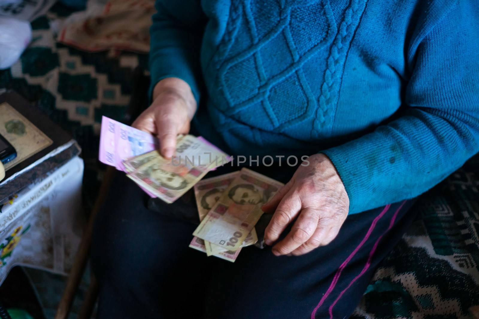 Old poor gray hair woman holds Ukrainian paper money in her hands. Woman is sad. Poor life in village. Old age not good. Low-light photo.