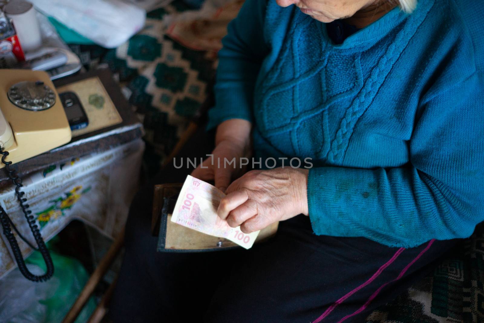 Old poor gray hair woman holds Ukrainian hryvna money in her hands. Woman is sad. Poor life in village. Old age not good. Low-light photo. by alexsdriver