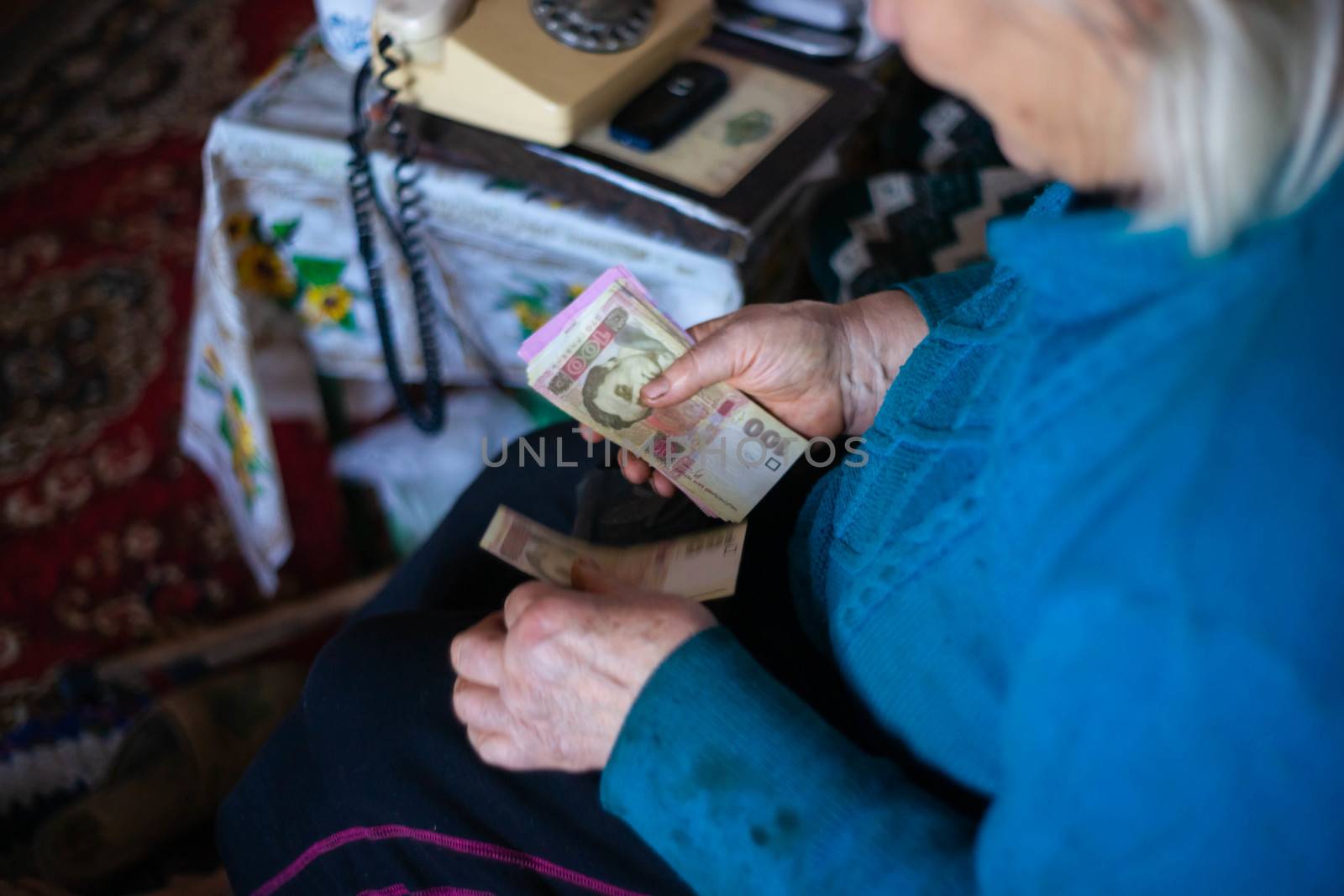 Old poor gray hair woman holds Ukrainian paper money in her hands. Woman is sad. Poor life in village. Old age not good. Low-light photo.