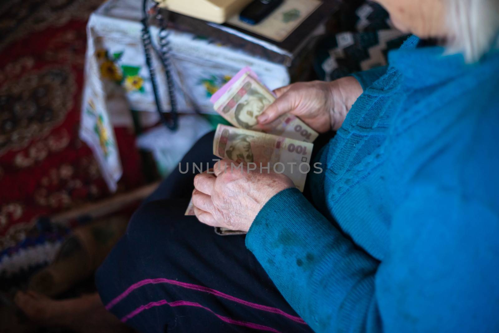 Old poor gray hair woman holds Ukrainian paper money in her hands. Woman is sad. Poor life in village. Old age not good. Low-light photo. by alexsdriver