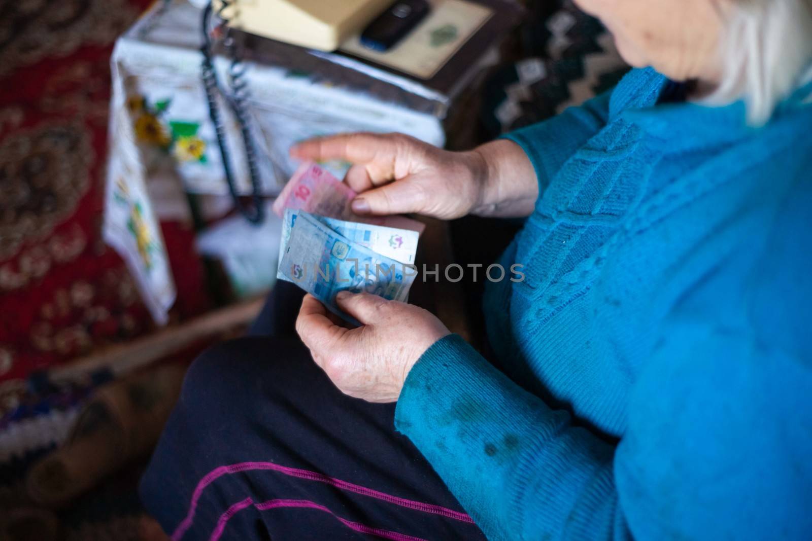 Old poor gray hair woman holds Ukrainian paper money in her hands. Woman is sad. Poor life in village. Old age not good. Low-light photo. by alexsdriver