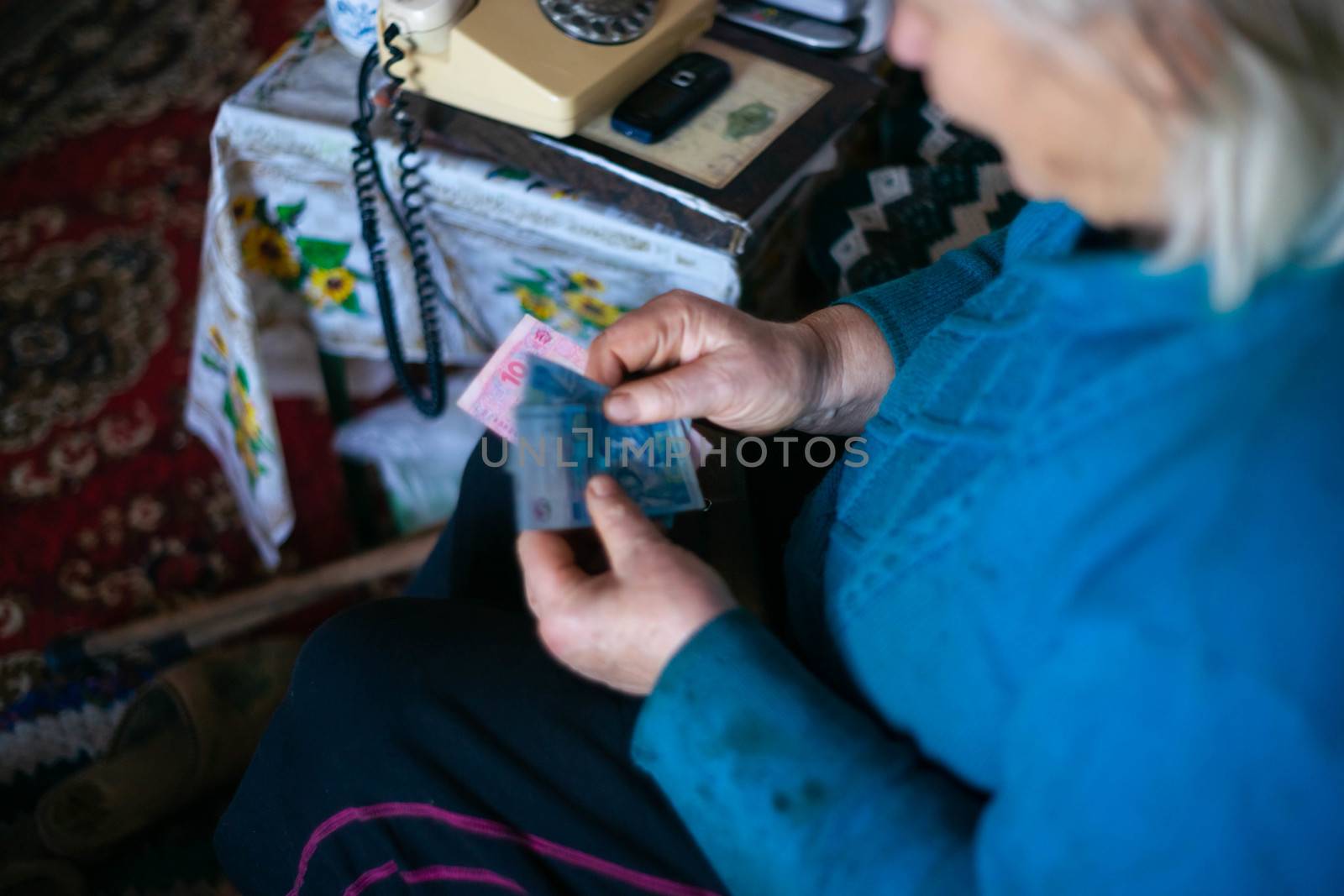 Old poor gray hair woman holds Ukrainian paper money in her hands. Woman is sad. Poor life in village. Old age not good. Low-light photo. by alexsdriver