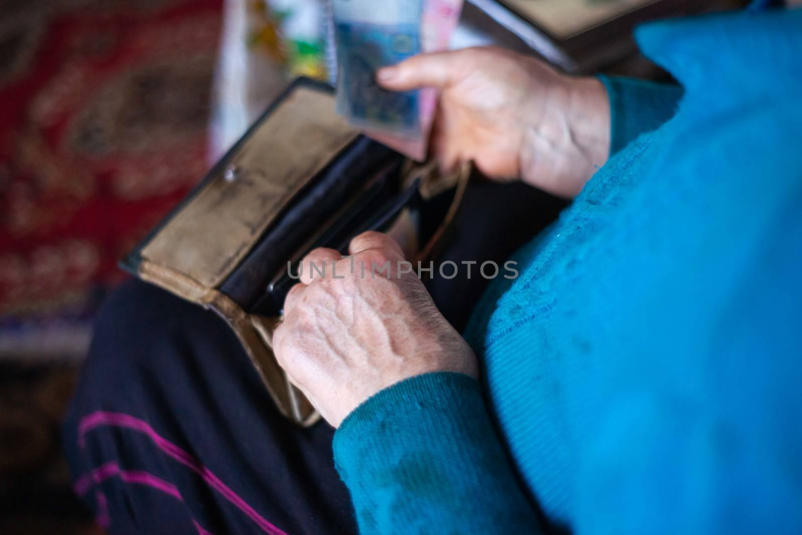 Old poor gray hair woman putting paper Ukrainian money in her vintage leather wallet by her hands. Woman is sad. Poor life in village. Old age not good. Low-light photo.