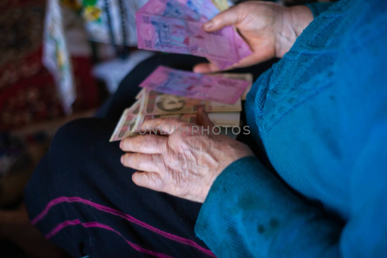 Old poor gray hair woman holds Ukrainian paper money in her hands. Woman is sad. Poor life in village. Old age not good. Low-light photo. by alexsdriver