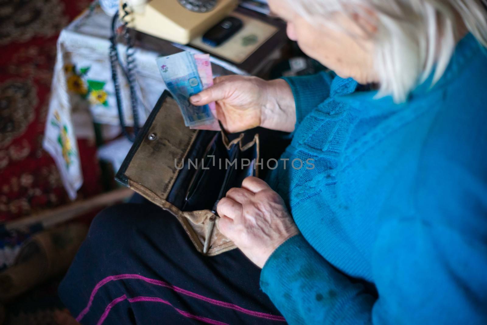 Old poor gray hair woman putting paper Ukrainian money in her vintage leather wallet by her hands. Woman is sad. Poor life in village. Old age not good. Low-light photo. by alexsdriver