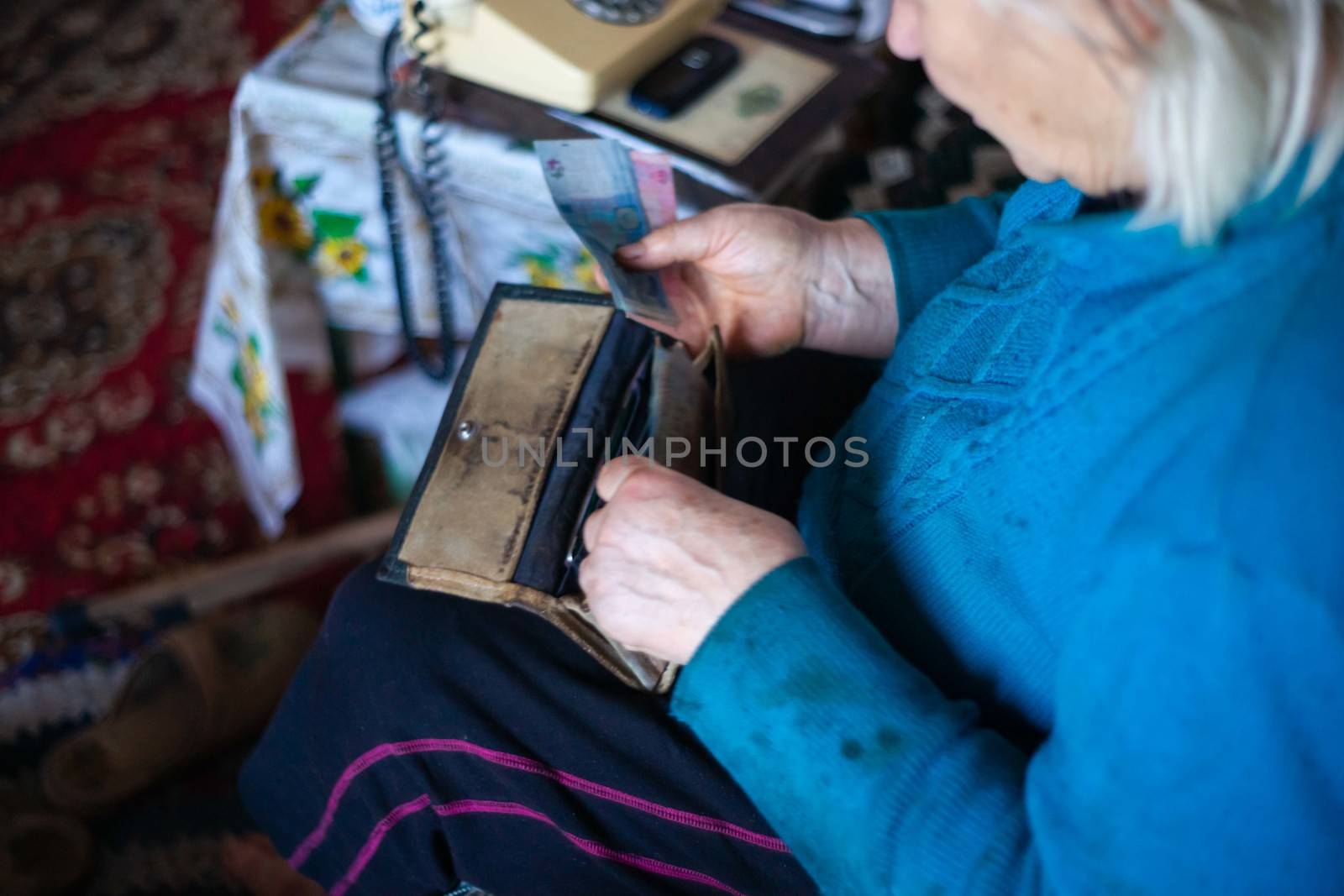 Old poor gray hair woman putting paper Ukrainian money in her vintage leather wallet by her hands. Woman is sad. Poor life in village. Old age not good. Low-light photo. by alexsdriver