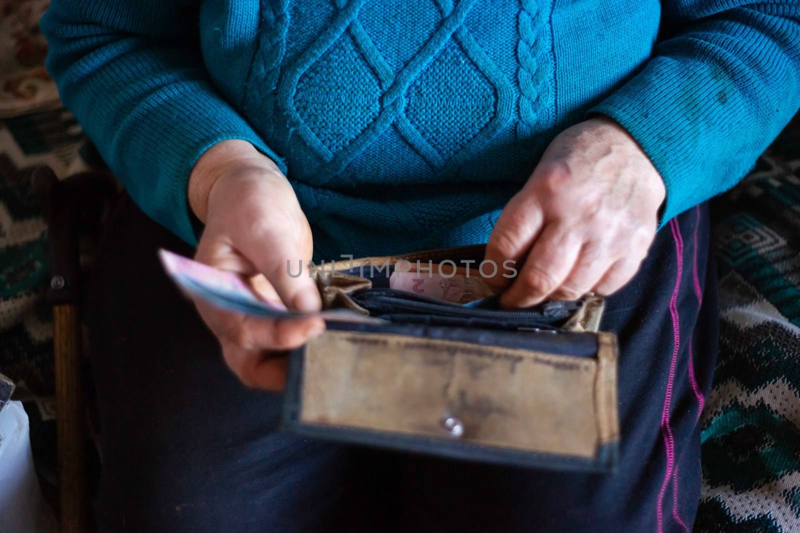 Old poor gray hair woman putting paper Ukrainian money in her vintage leather wallet by her hands. Woman is sad. Poor life in village. Old age not good. Low-light photo. by alexsdriver