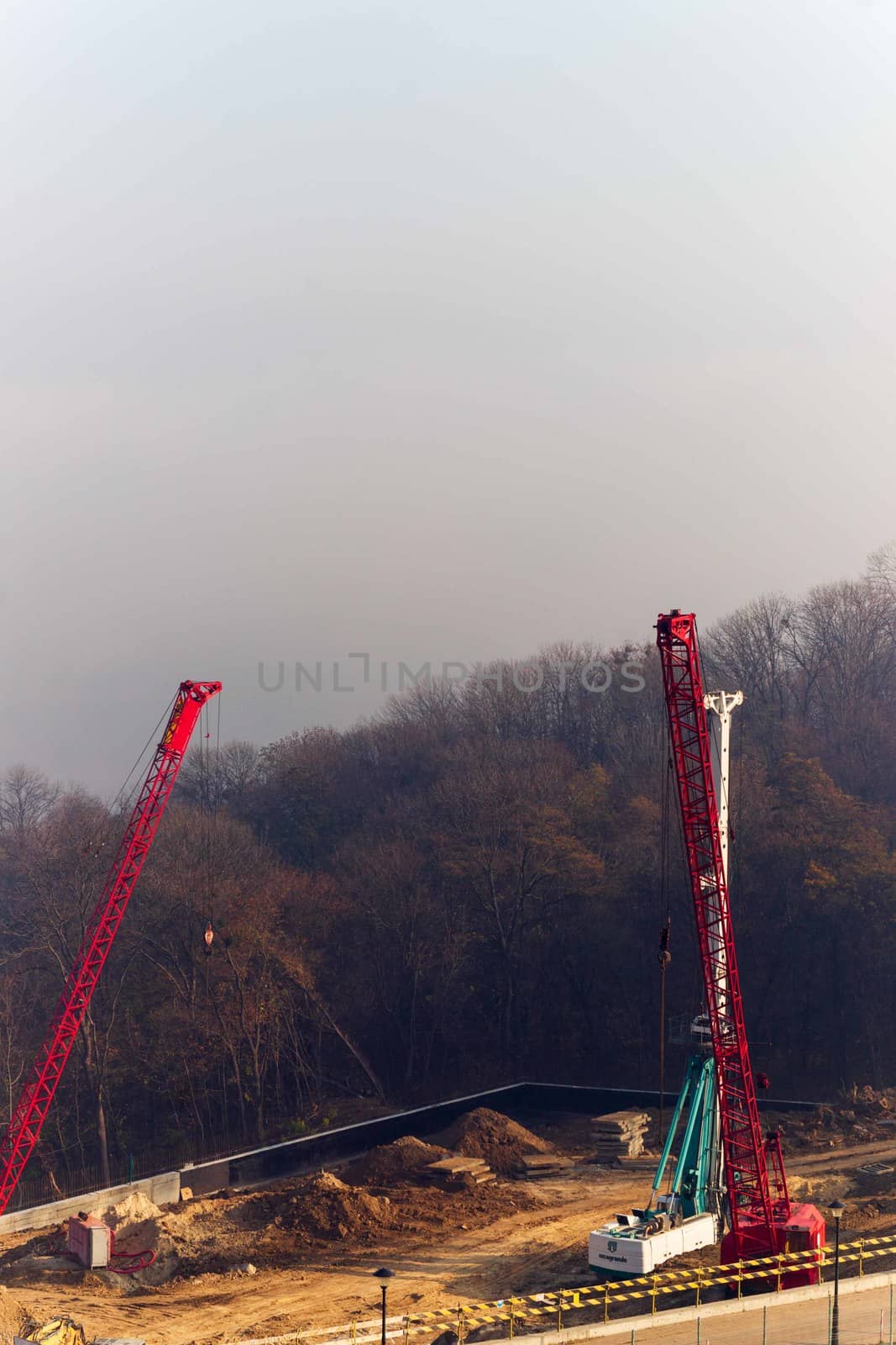 Construction process with construction automotive cranes with red and white gibbets. Autumn fog and river bridge  on blurred background. Forest around cranes.