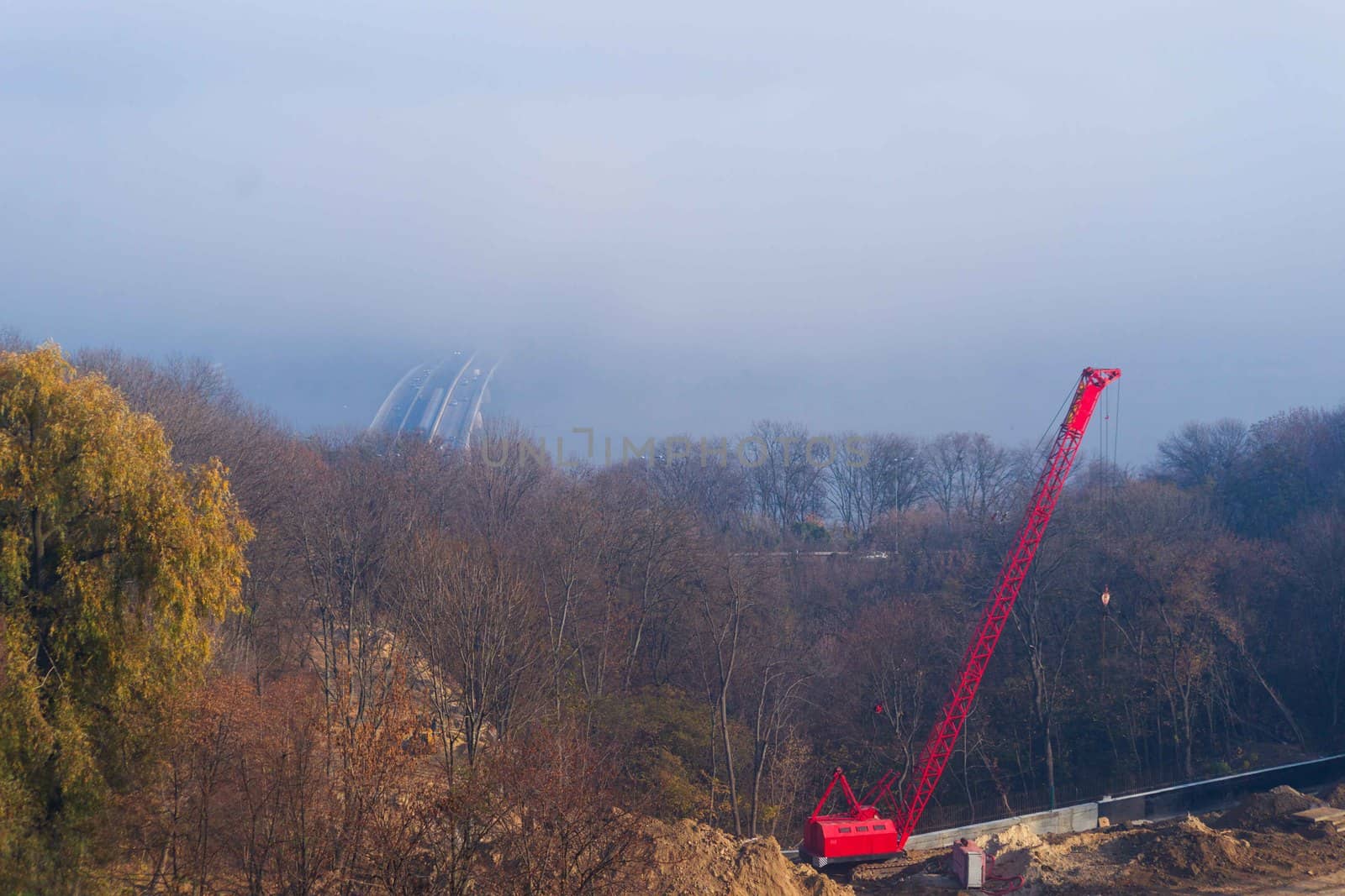 Construction process with construction automotive cranes with red  gibbet. Autumn fog and river bridge with subway train on blurred background. Forest around cranes.