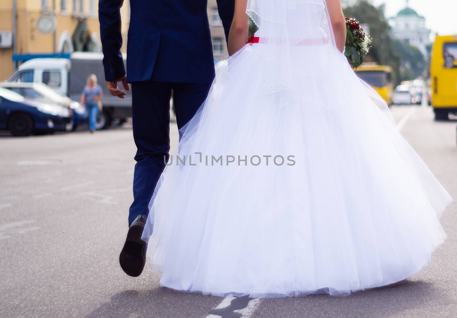 The bride and groom hold  hands themselves while they walking on the road in city. Wedding in detail.
