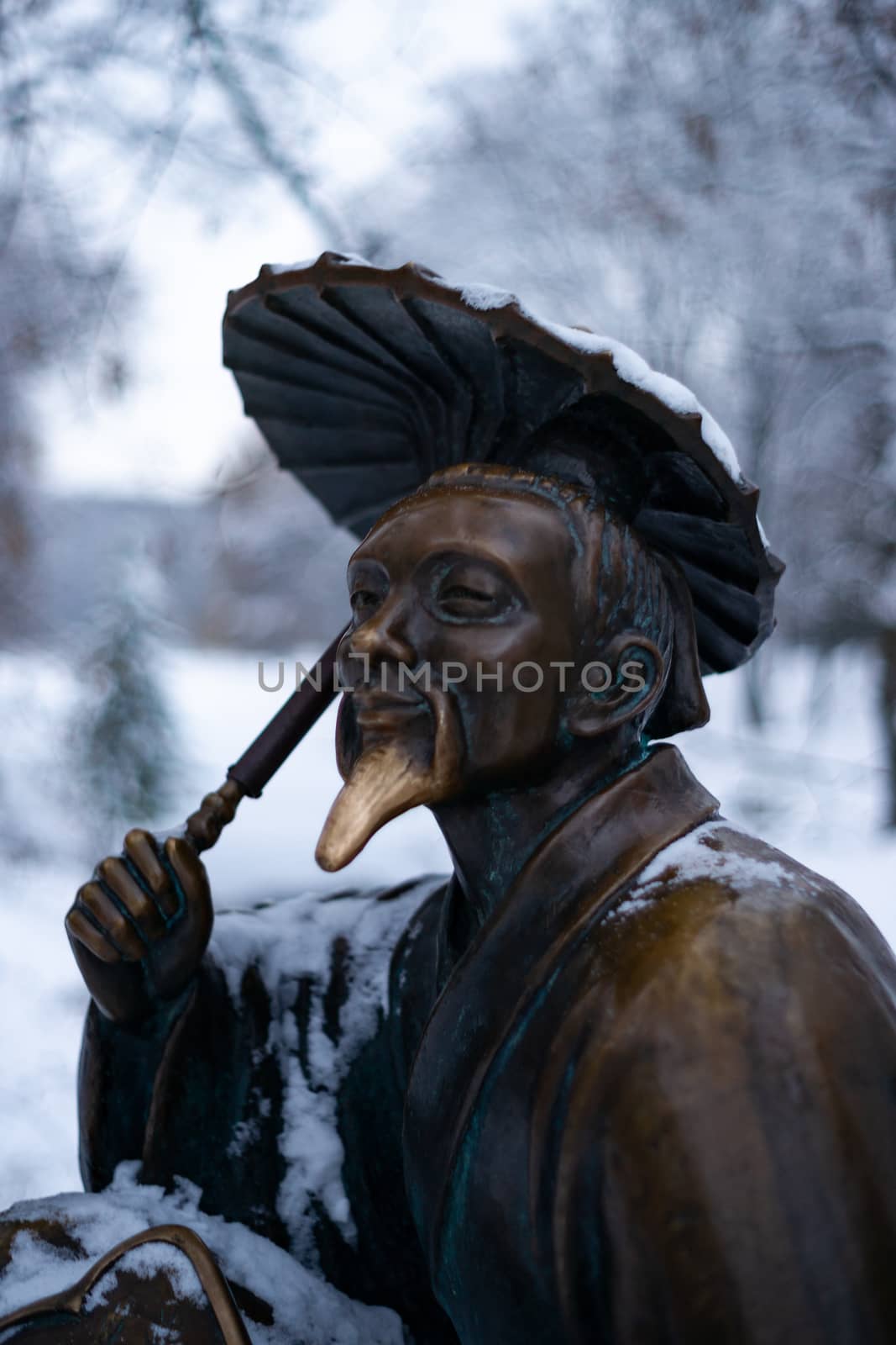 Bronze monument of a chinese old man with umbrella in winter snow park. Closeup with blurred background.