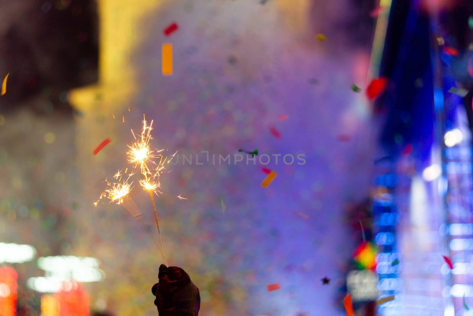 New Year celebration on main square. A lot of smoke or fog, lights, salute and confetti. Blurred colorful background. Bengali fire in hand with glove on foreground. Concert scene with blue light at right side of frame.