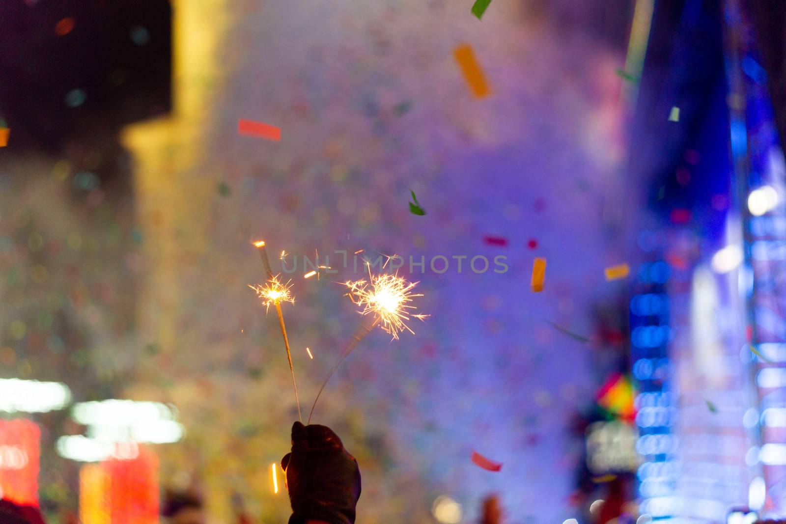 New Year celebration on main square. A lot of smoke or fog, lights, salute and confetti. Blurred colorful background. Bengali fire in hand with glove on foreground. Concert scene with blue light at right side of frame.