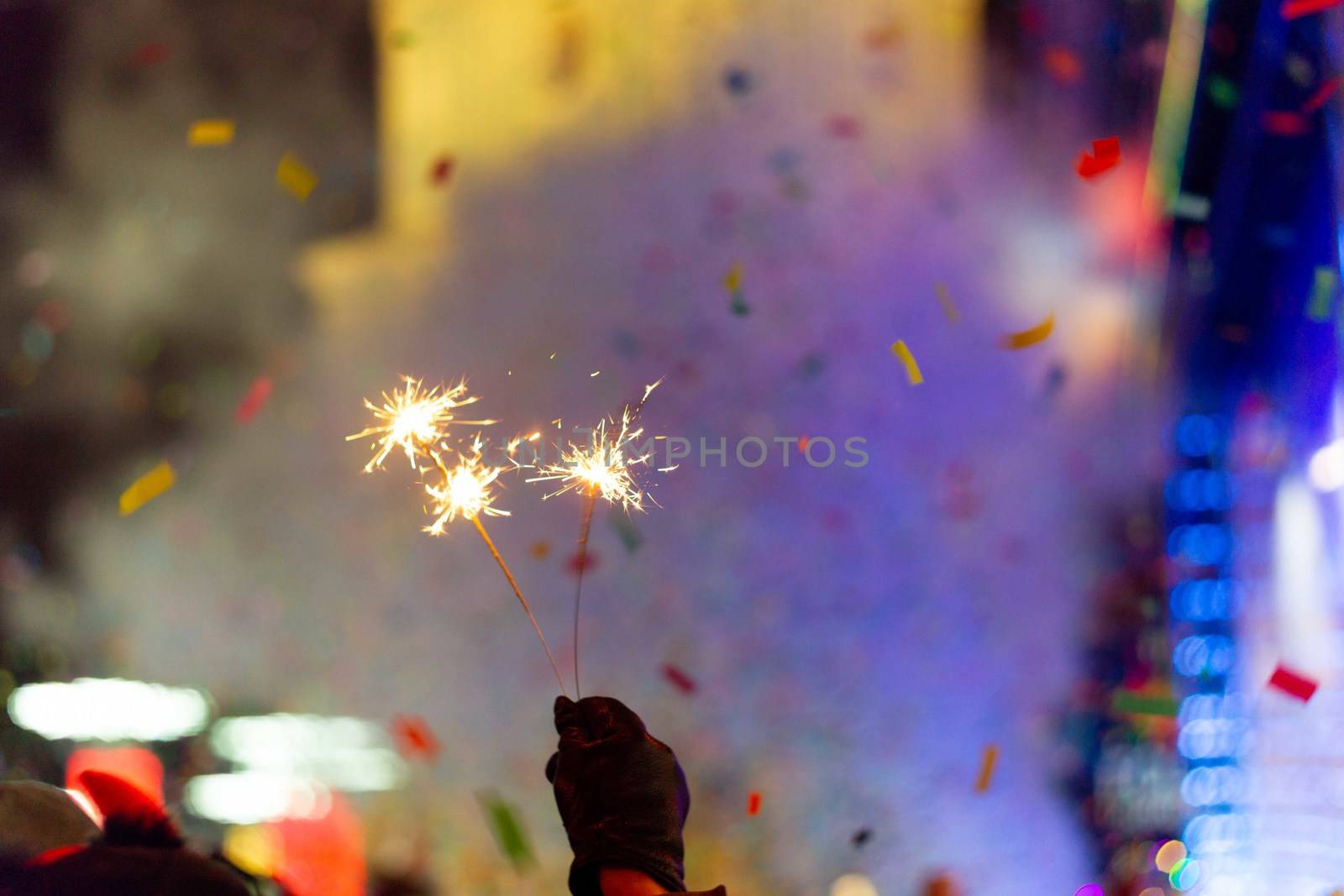 New Year celebration on main square. A lot of smoke or fog, lights, salute and confetti. Blurred colorful background. Bengali fire in hand with glove on foreground. Concert scene with blue light at right side of frame.