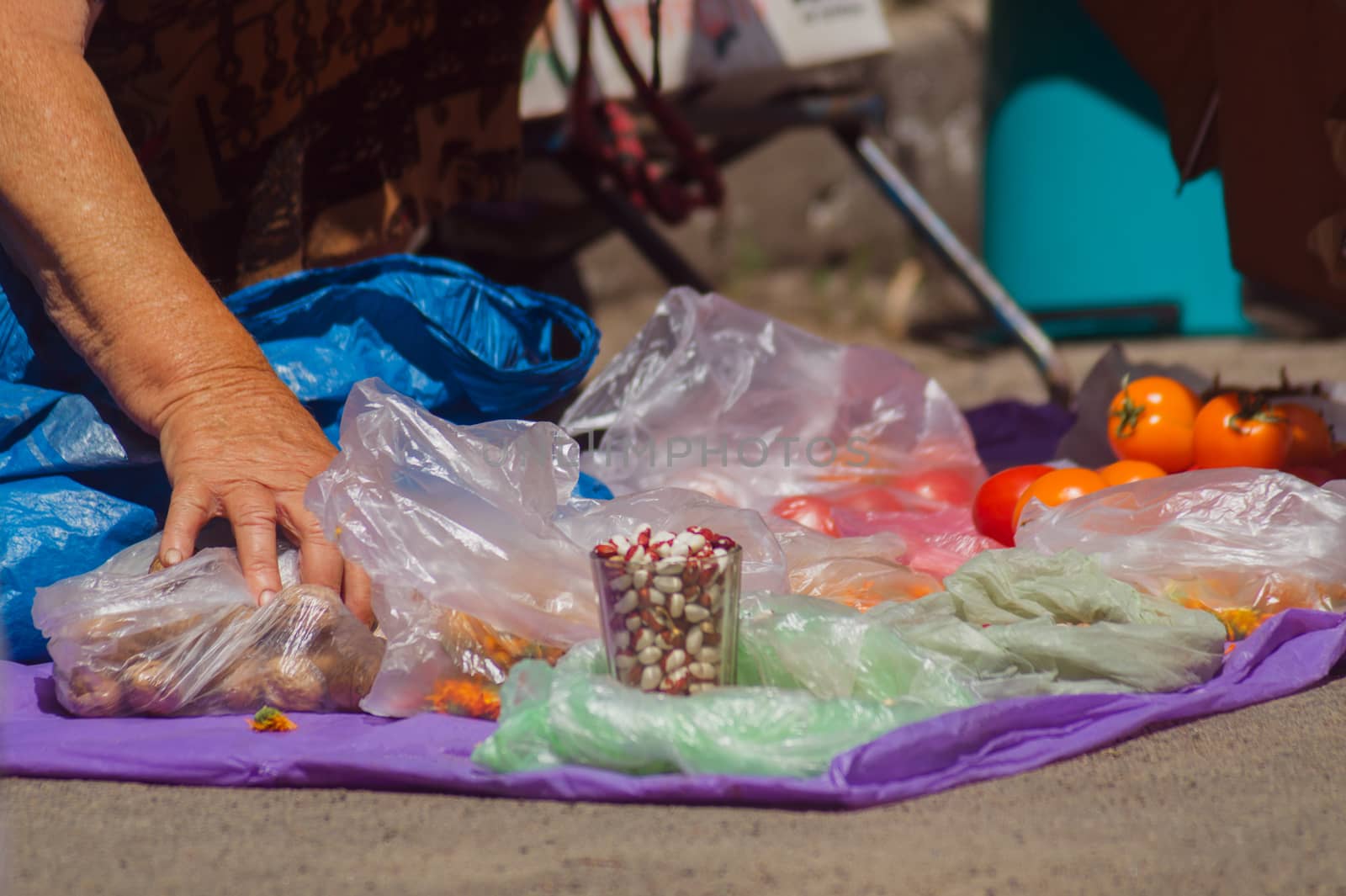 Street mart on asphalt. Old woman holds packet with potato. In foreground is a glass of beans.