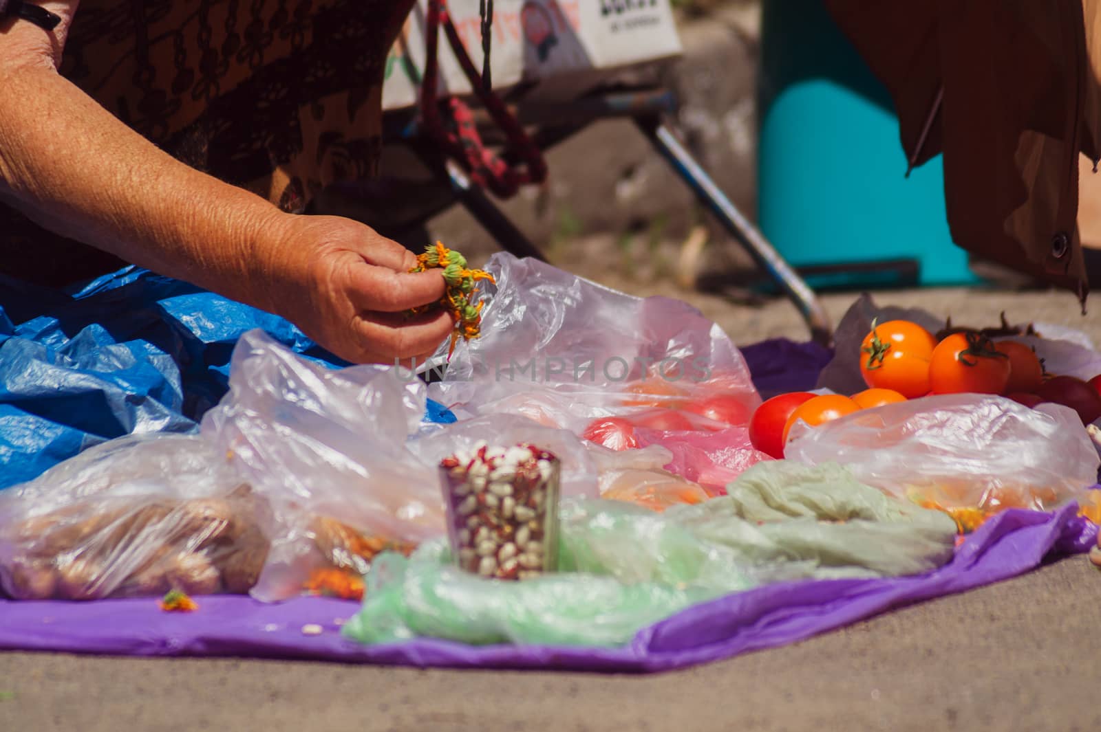 Street mart on asphalt. Old woman holds cutted flowers in her hand. In  foreground is a glass of beans.