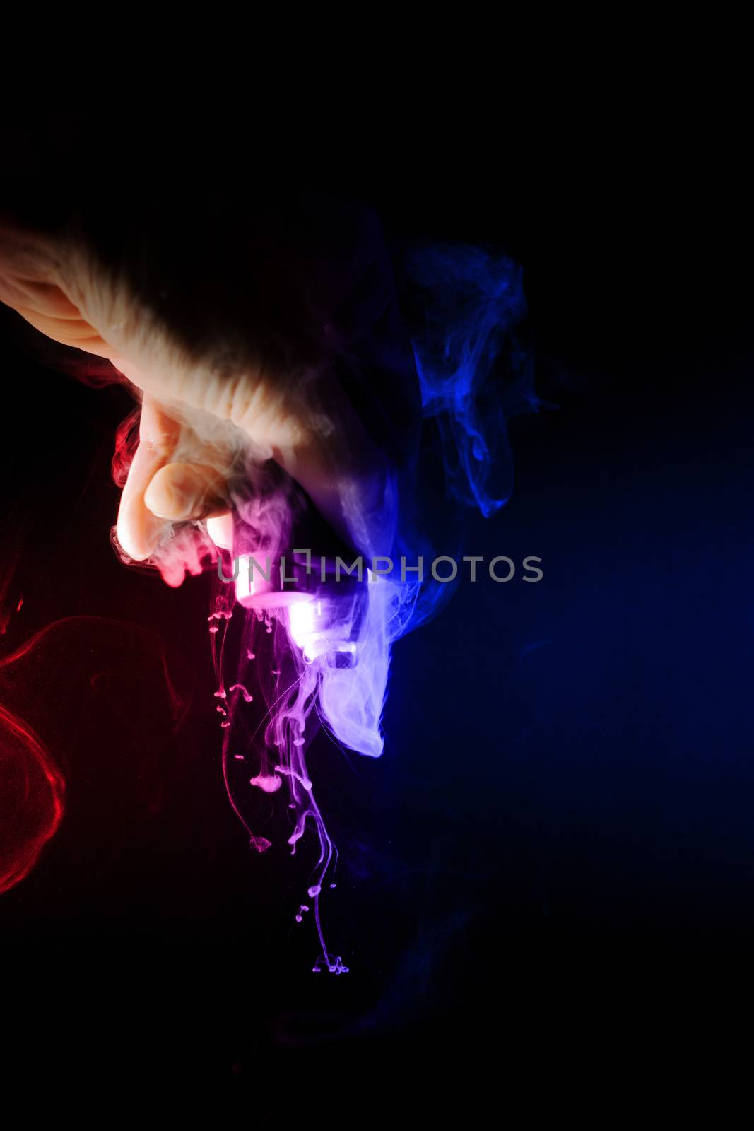Male hand holds vape upside down. Vape clouds splash from coil on black background. Fog is blue and red colour. Stock isolated smoke with spray boiling glycerine.