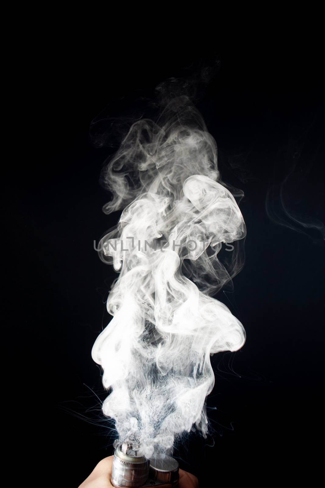 Man hand holds vape. Vape clouds splash from coil on black background. Fog is white. Stock isolated white smoke with spray boiling glycerine around.