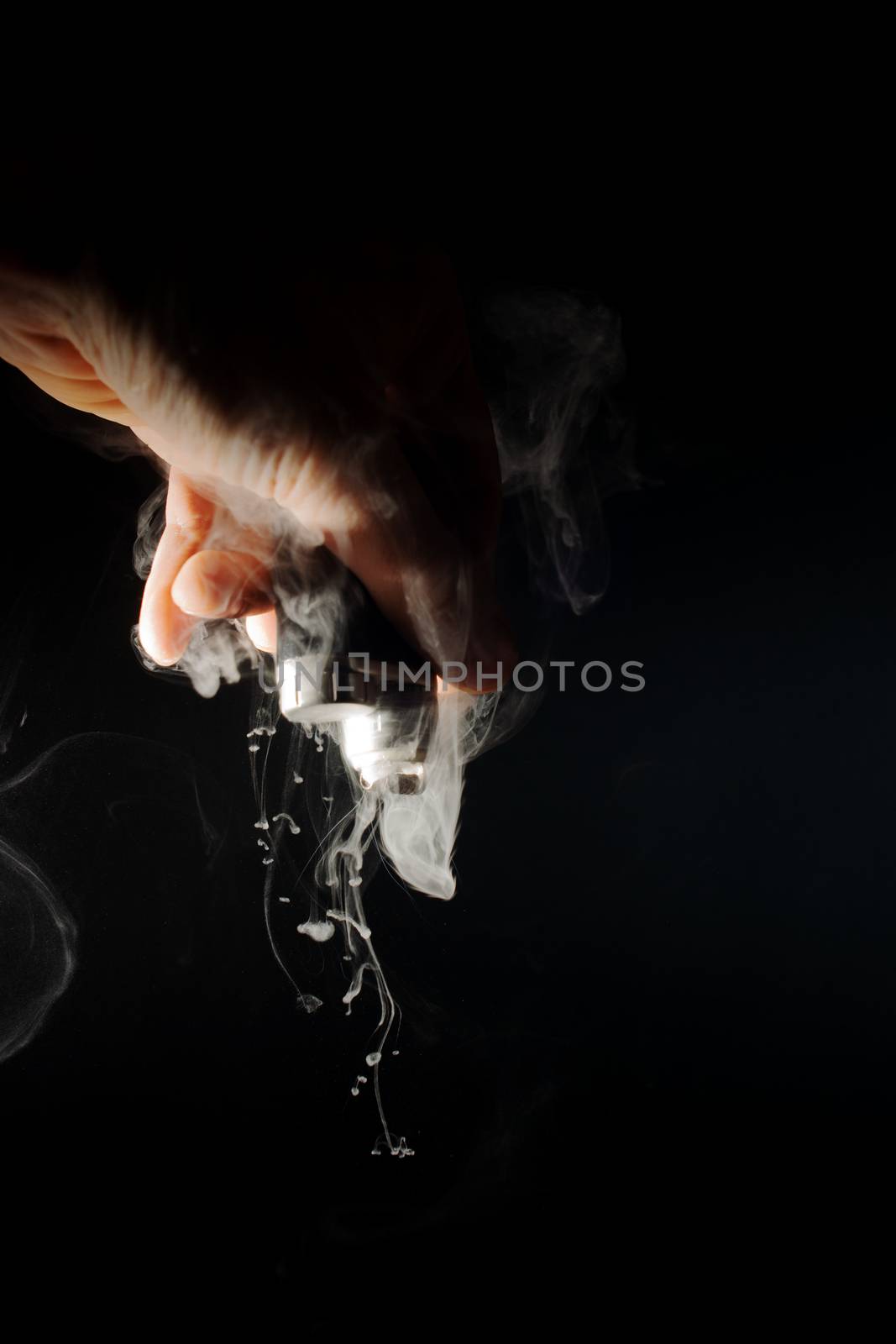 Male hand holds vape upside down. Vape clouds splash from coil. Fog is white. Stock isolated white smoke with spray boiling glycerine on black ground.