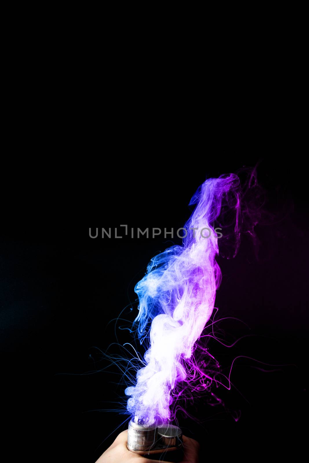 Young man holds vape in his hand. Vape clouds splash from vape coil on black background. Fog is dual color — blue and purple. Stock isolated colorful smoke with spray boiling glycerine.