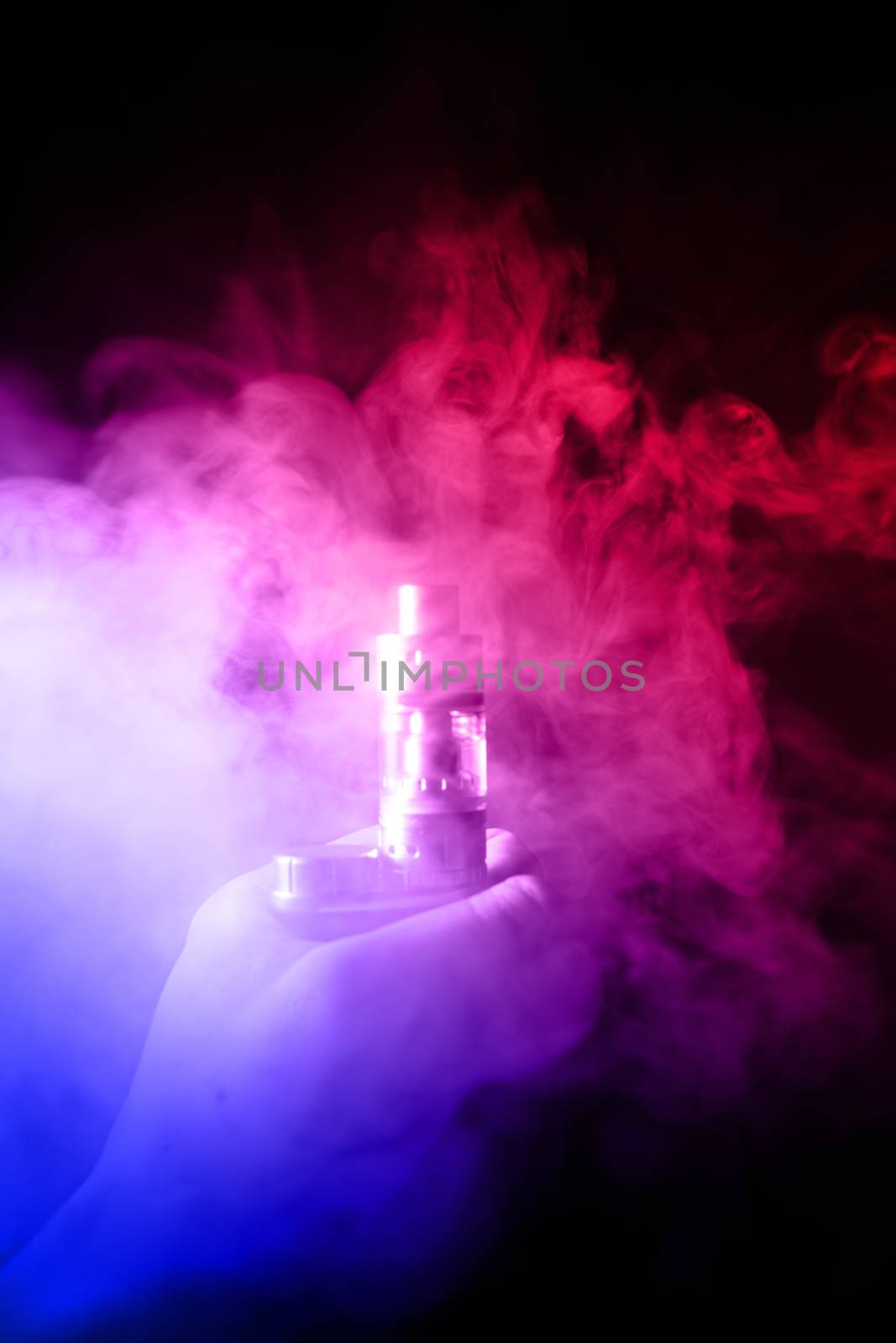 Male hand holds vape with atomiser in thick vape fog or smoke. Vape clouds around hand with vape on black background. Fog is dual color — blue and red. Stock isolated colorful smoke with spray glycerine.