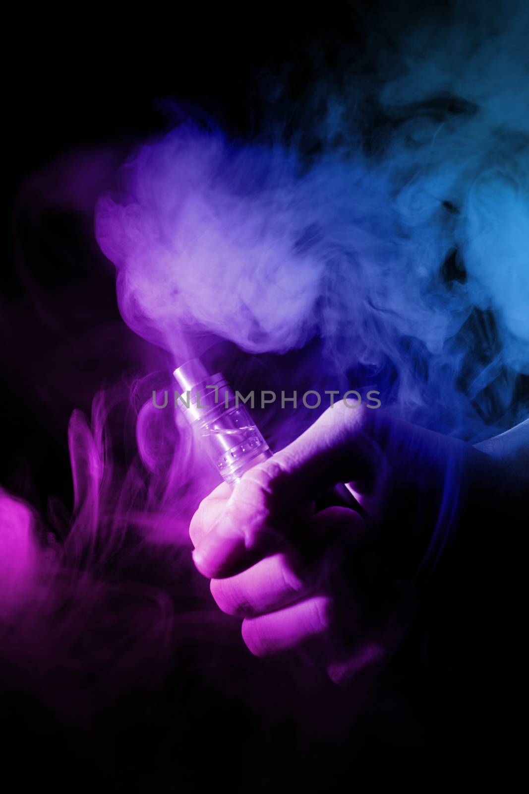 Male hand holds vape in thin vape fog or smoke. Vape clouds around hand with vape on black background. Light is dual color — blue and purple. Stock isolated colorful smoke with spray glycerine.