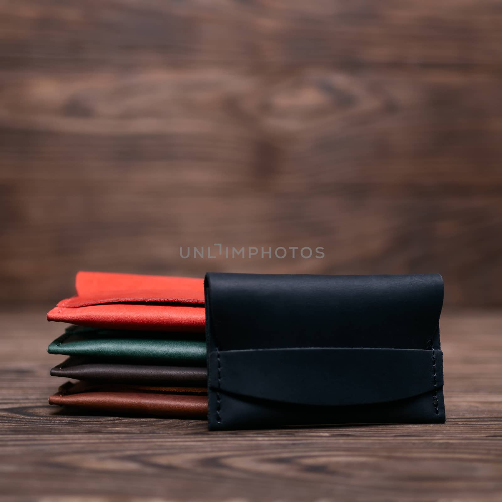 Flat lay photo of five different colour handmade leather one pocket cardholders. Black cardholder on foreground. Red, black, blown, ginger and green colors. Stock photo on wooden background.