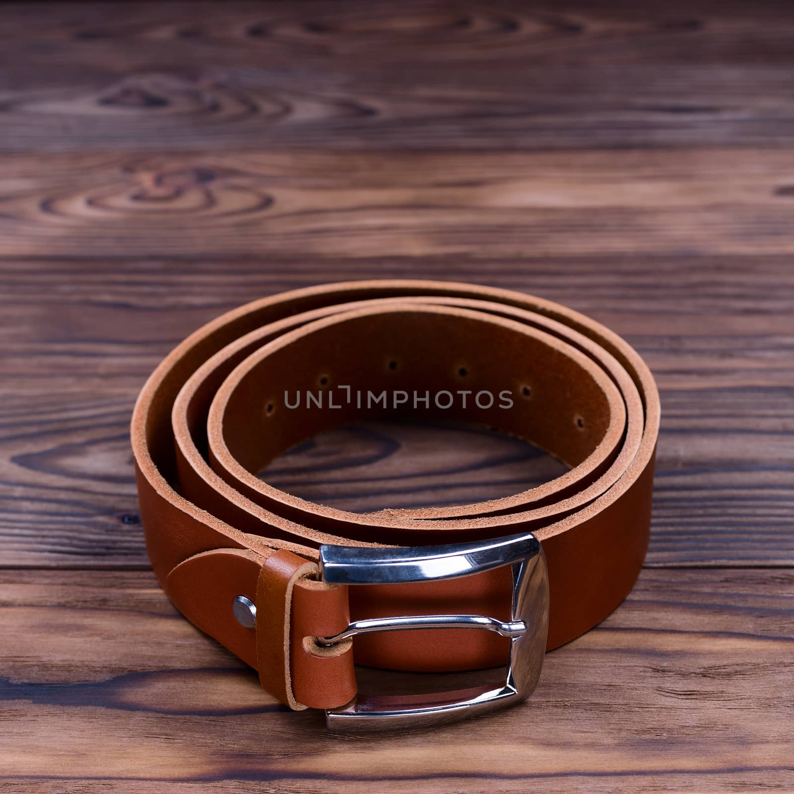 Ginger color handmade belt lies on textured wooden background. The belt is twisted into a ring. Closeup side view. Stock photo of businessman accessories.