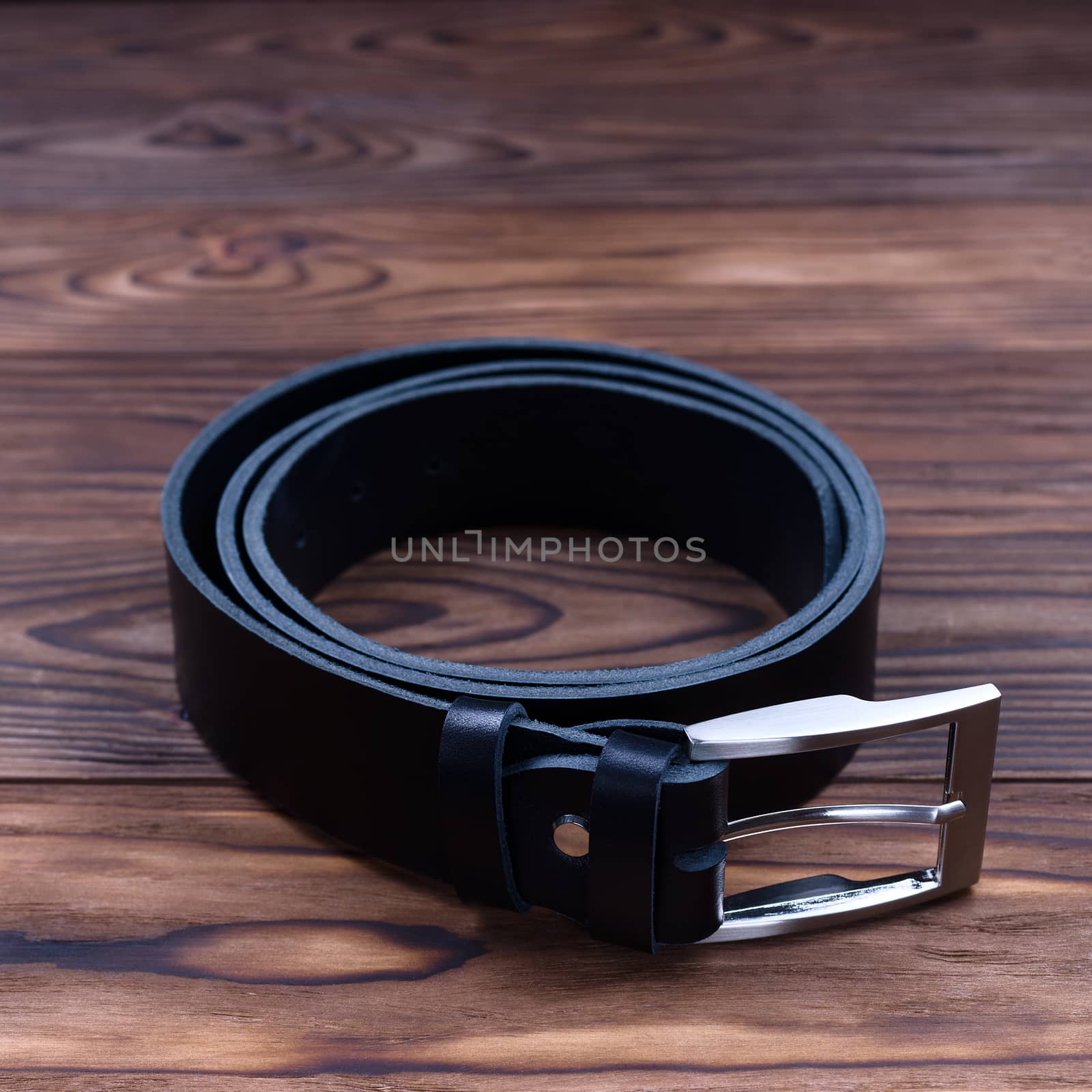 Black color handmade belt lies on textured wooden background. The belt is twisted into a ring. Closeup side view. Stock photo of businessman accessories.