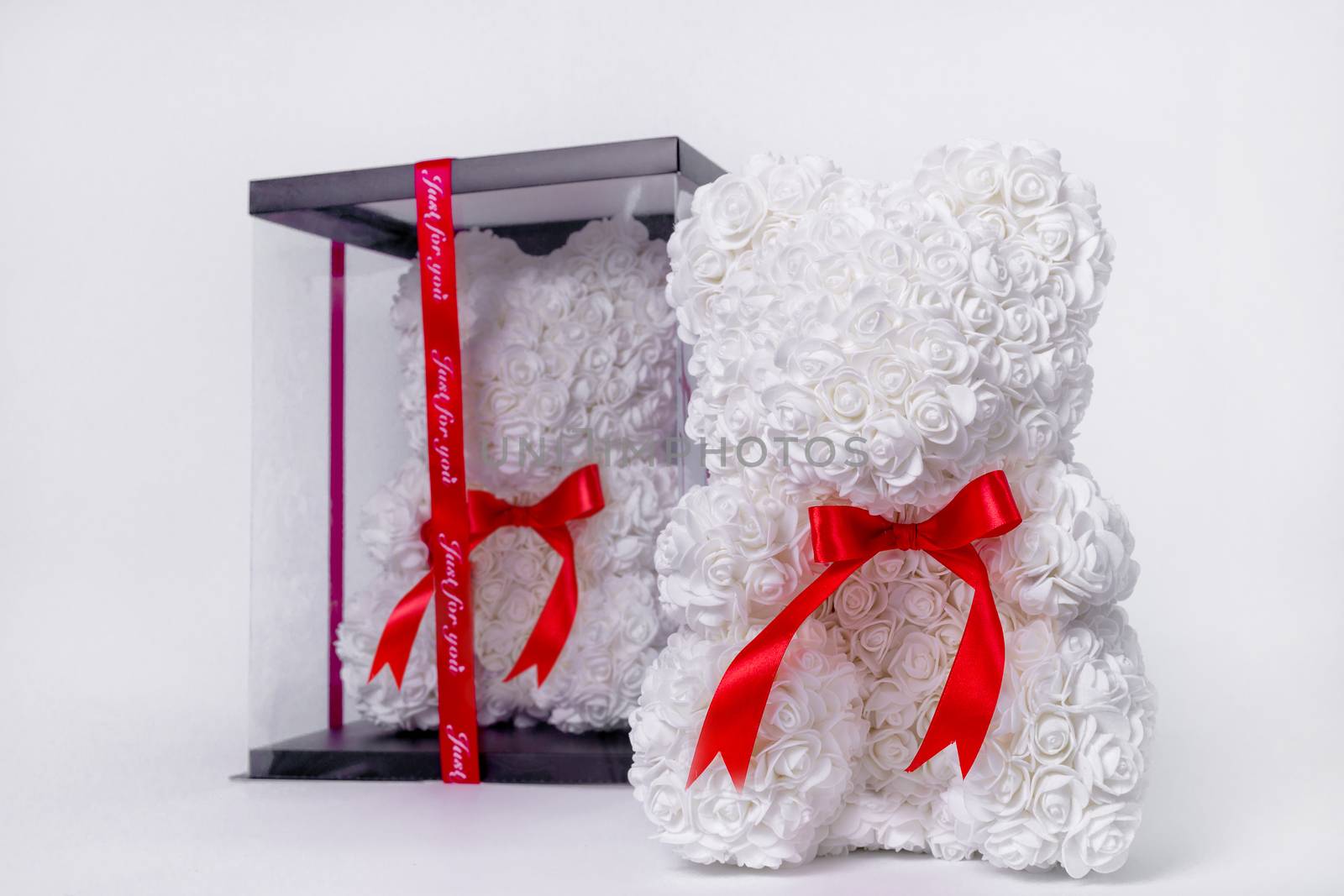 White teddy bear toy of foamirane roses. The same teddy in clear box with black paper cover on background. Red stripe on teddy neck. Stock photo isolated on white background. Gift on holiday for women.