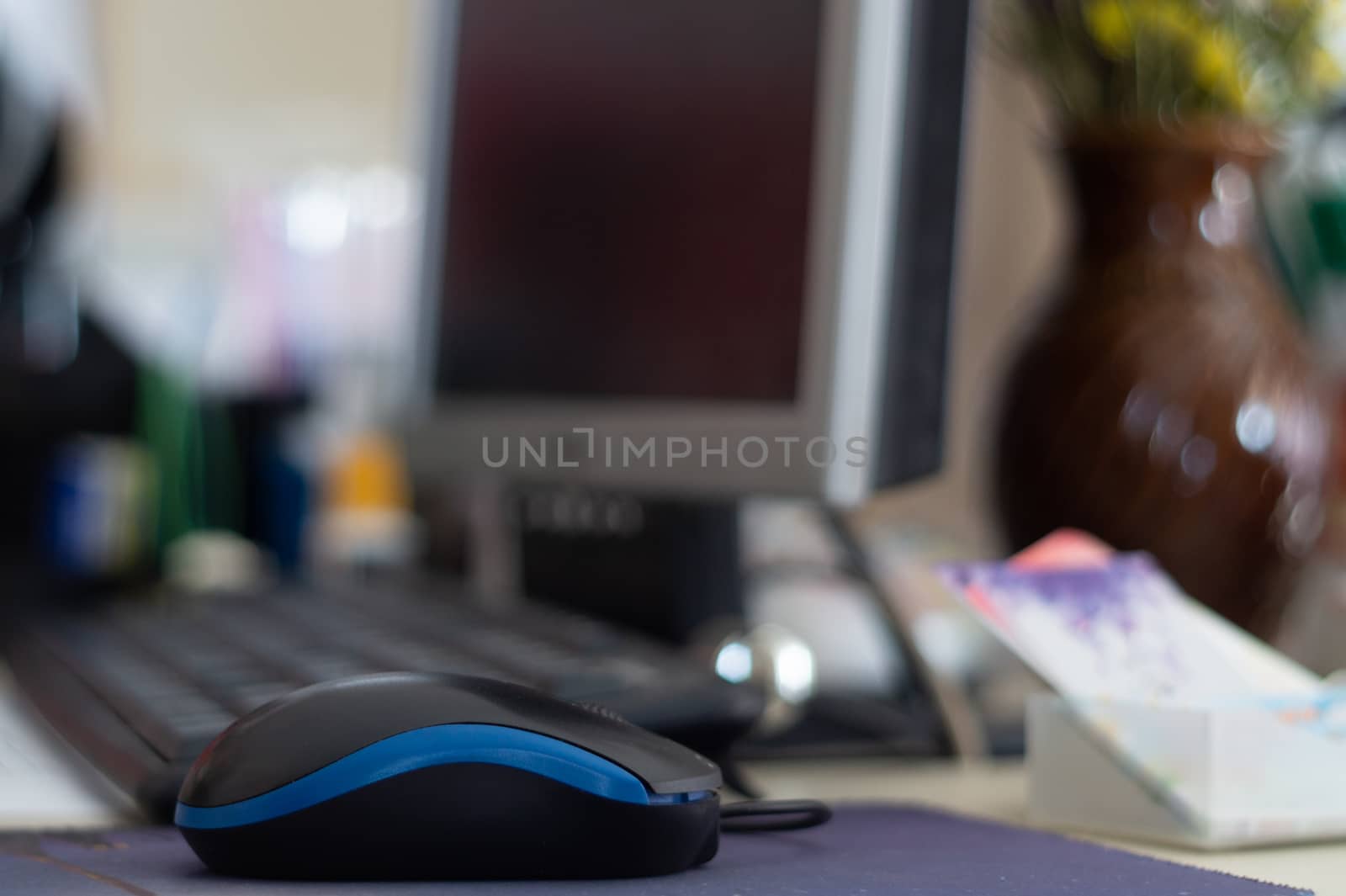 Small digital mouse on the table. On background is desktop monitor, bouquet of flowers and keyboard