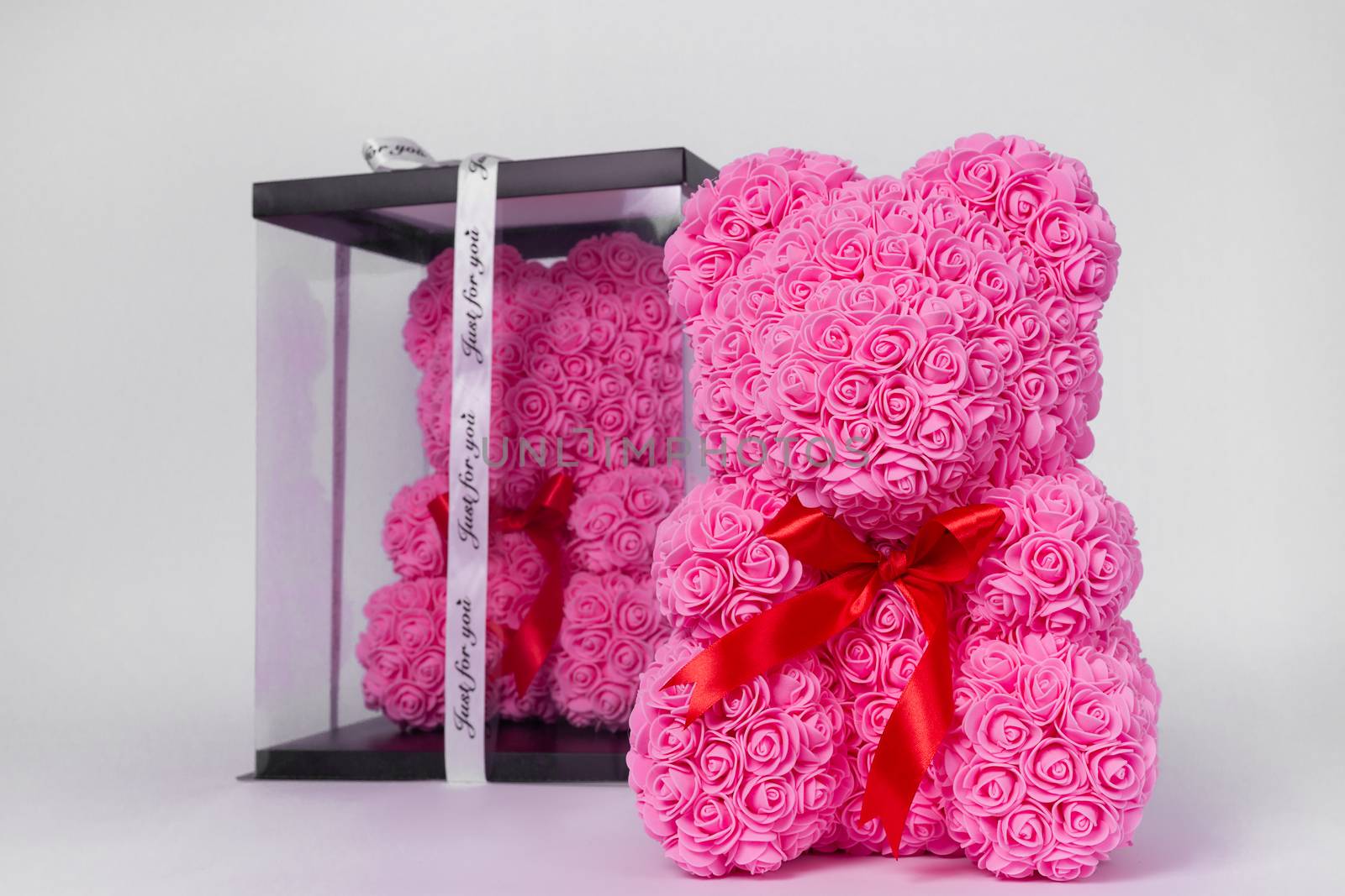 Pink teddy bear toy of foamirane roses. The same teddy in clear box with black paper cover on background. Red stripe on teddy neck. Stock photo isolated on white background. Gift on holiday for women.
