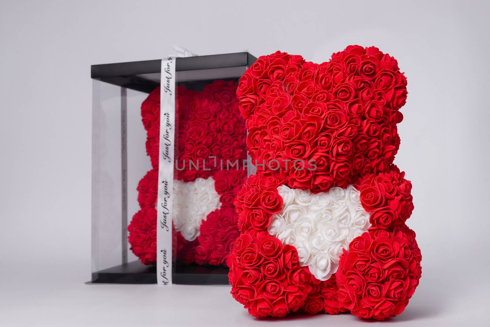 Red  teddy bear of foamirane roses. The same teddy in clear box with black paper cover on background. White heart in teddy paws. Stock photo isolated on white background.