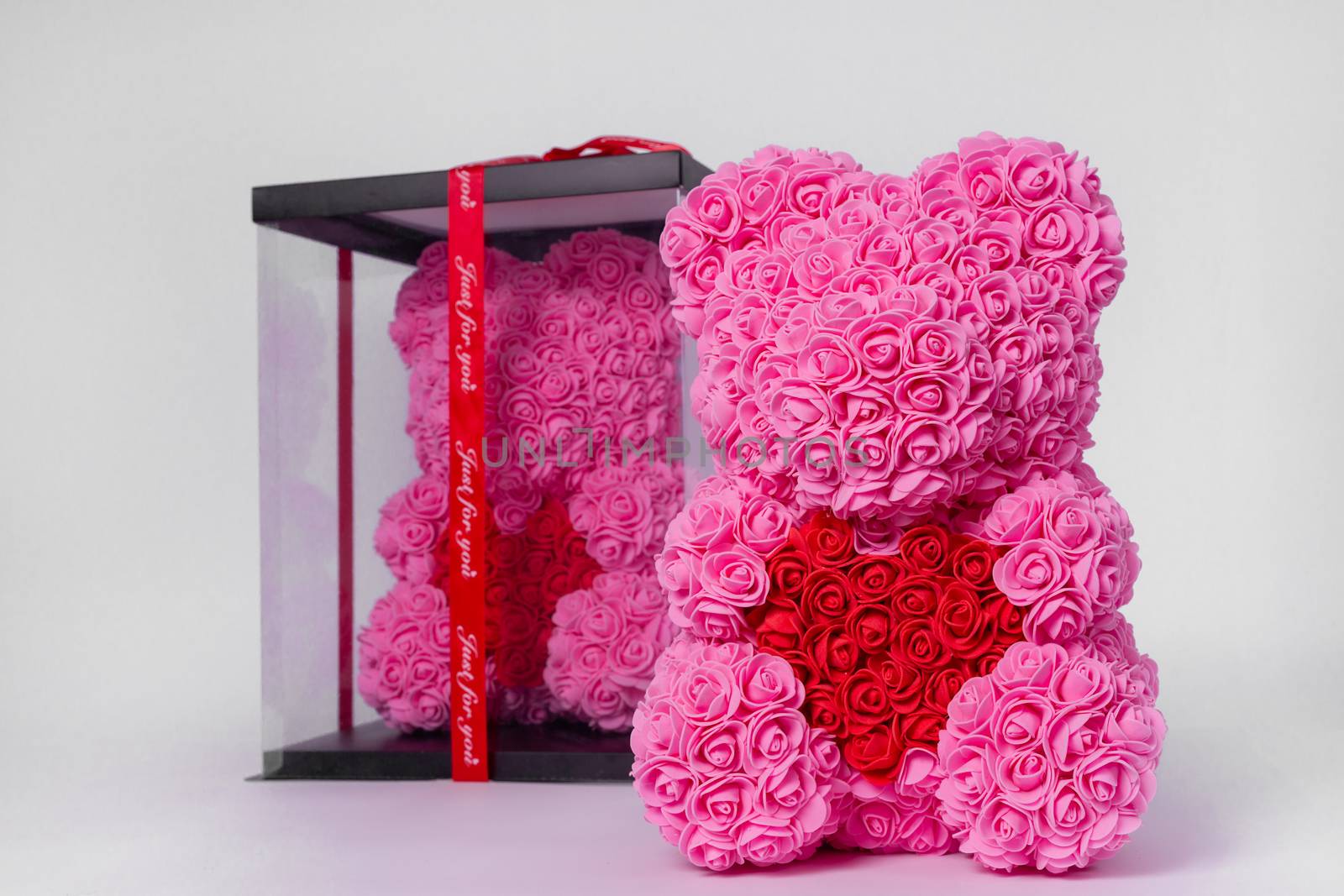 Pink  teddy bear toy of foamirane roses. The same teddy in clear box with black paper cover on background. Red heart in teddy paws. Stock photo isolated on white background.