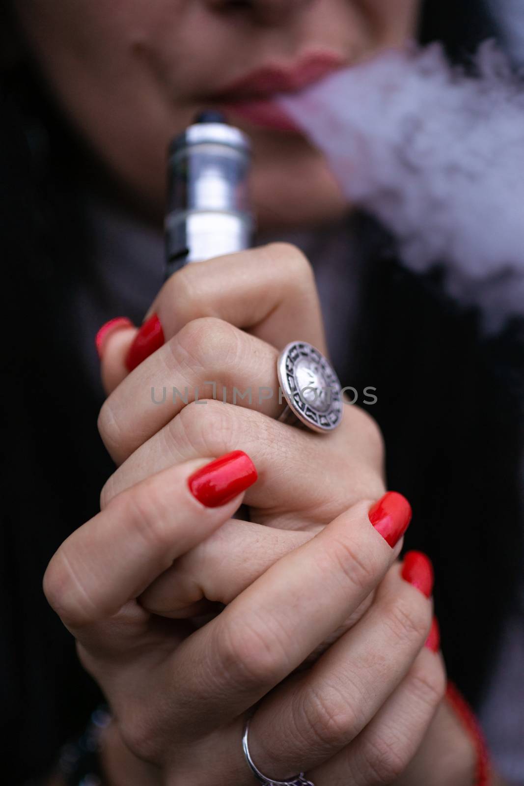 Caucasian woman with red nails manicure and antique ring on finger holds small vape. Smoking alternative vay. Woman exhales thick smoke. Life without cigarettes. Woman-vaper. Small e-cigarette.