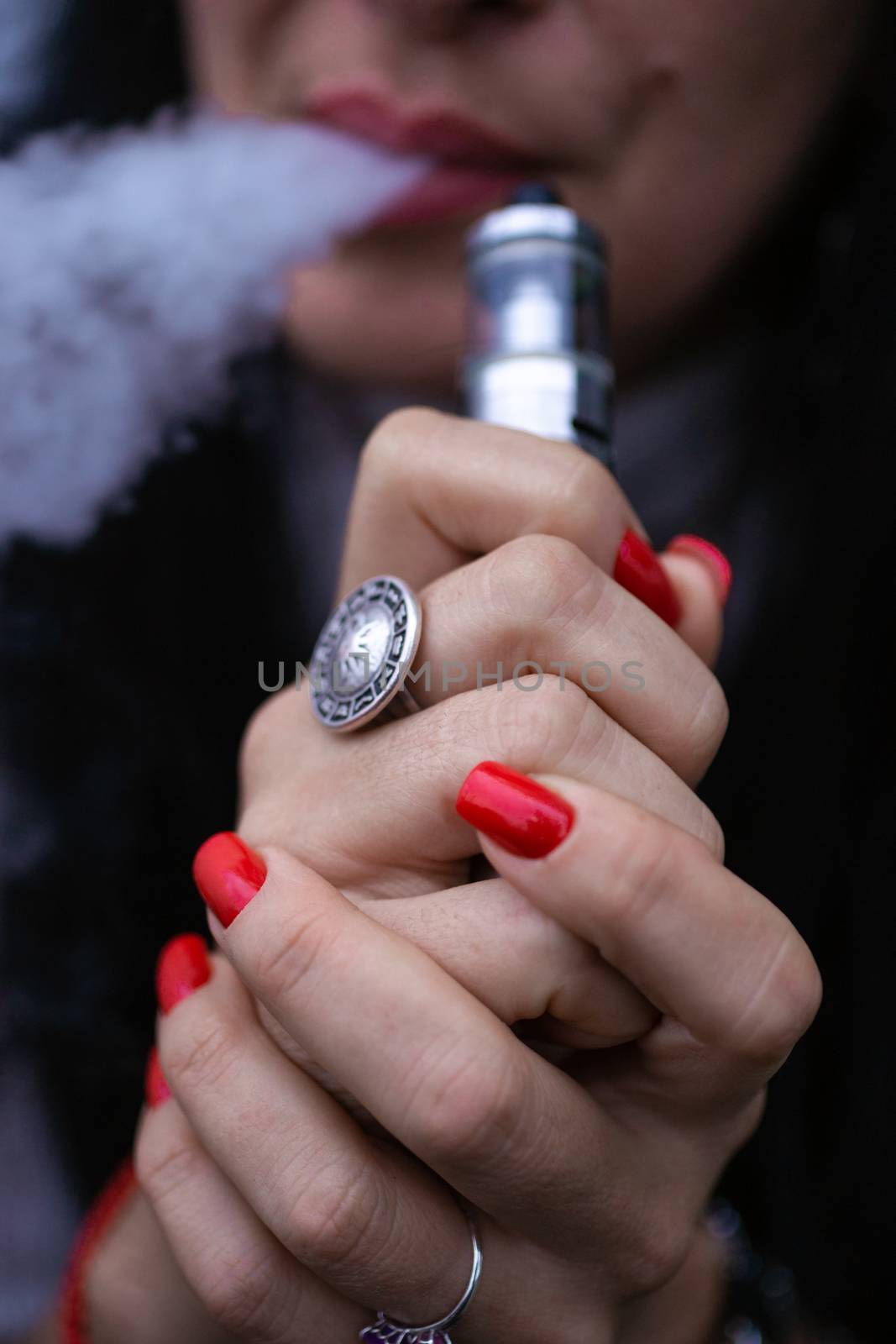 Caucasian woman with red nails manicure and antique ring on finger holds small vape. Smoking alternative vay. Woman exhales thick smoke. Life without cigarettes. Woman-vaper. Small e-cigarette.