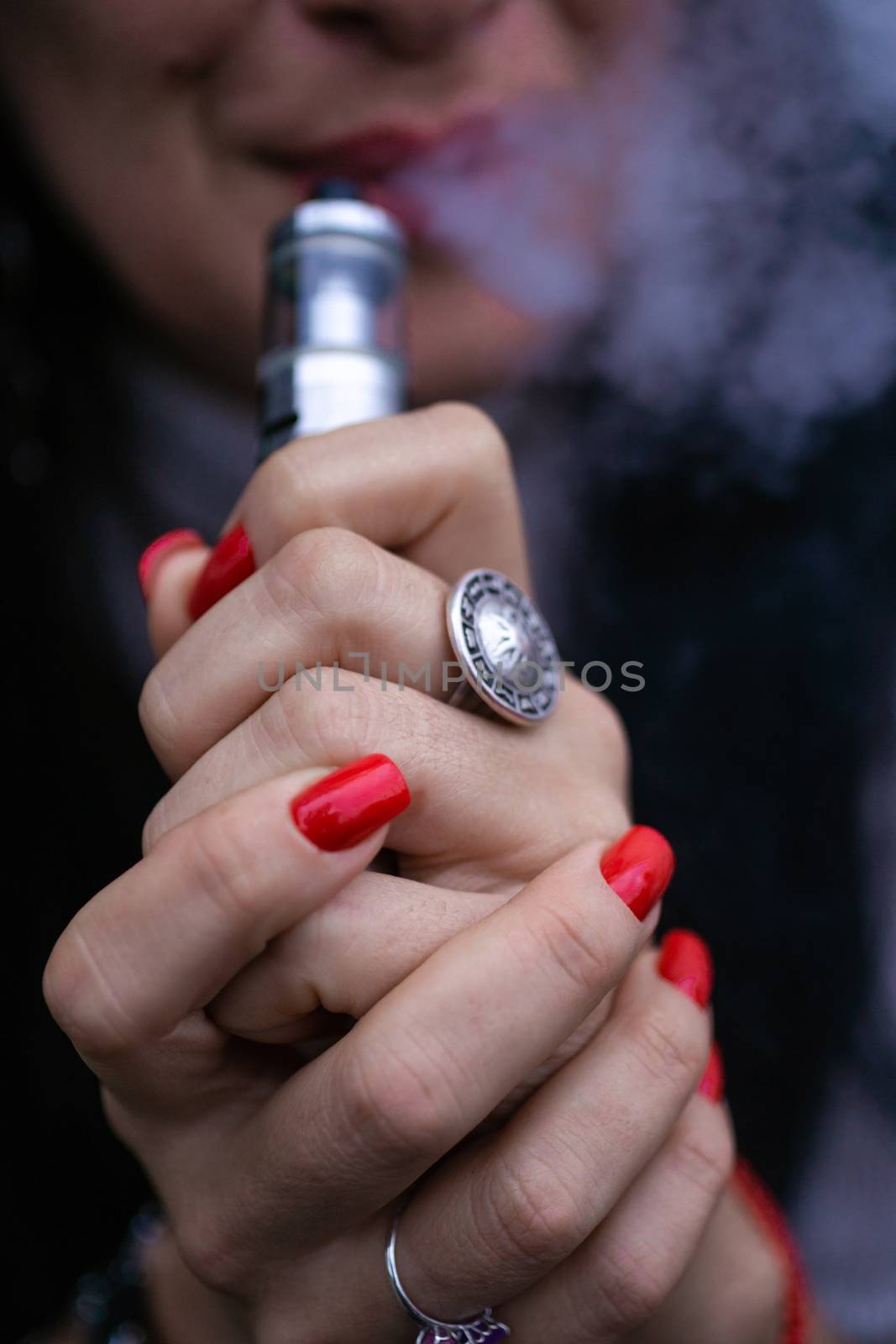 Caucasian woman with red nails manicure and antique ring on finger holds small vape. Smoking alternative vay. Woman exhales thick smoke. Life without cigarettes. Woman-vaper. Small e-cigarette. by alexsdriver