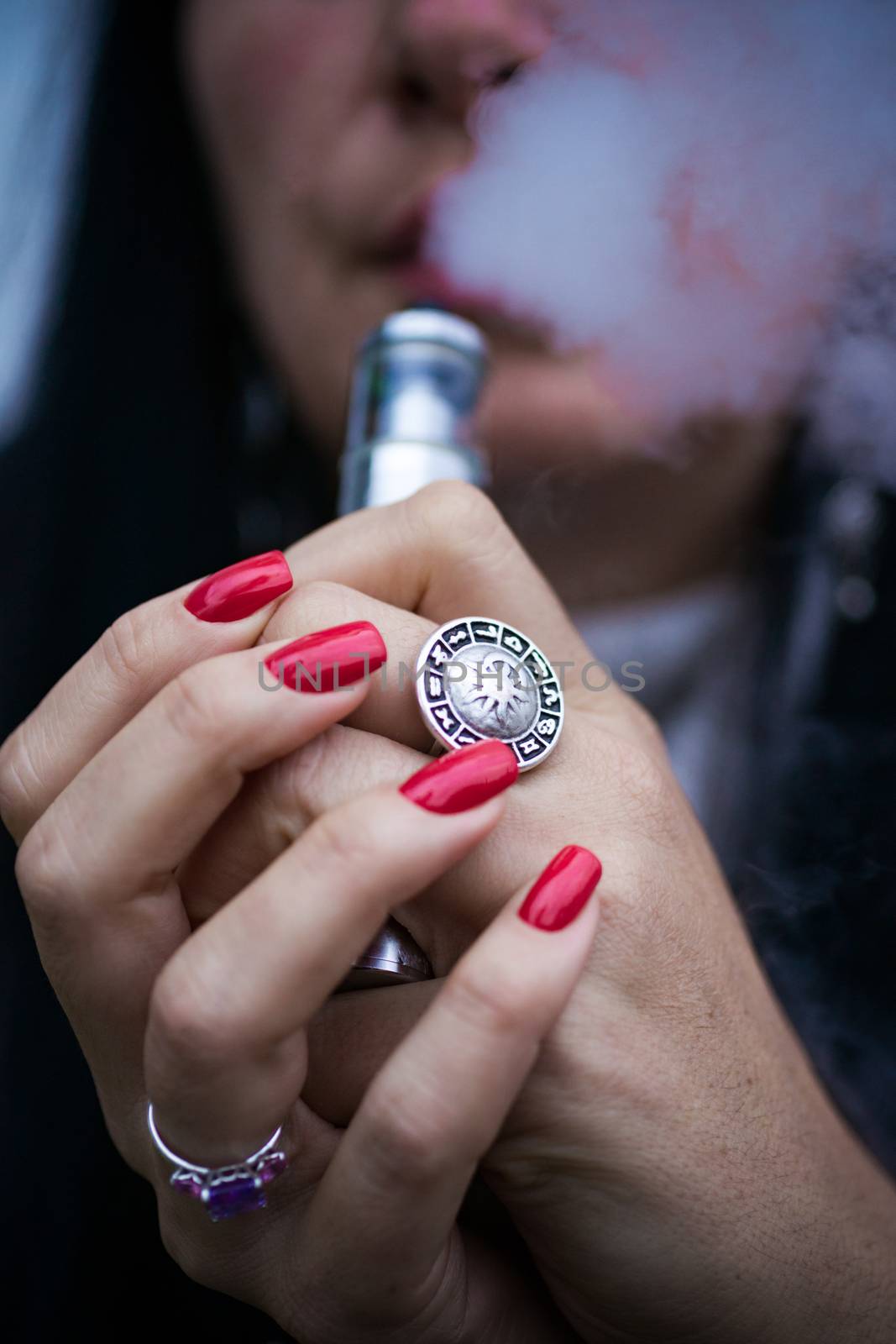 Caucasian woman with red nails manicure and antique ring on finger holds small vape. Smoking alternative vay. Woman exhales thick smoke. Life without cigarettes. Woman-vaper. Small e-cigarette.
