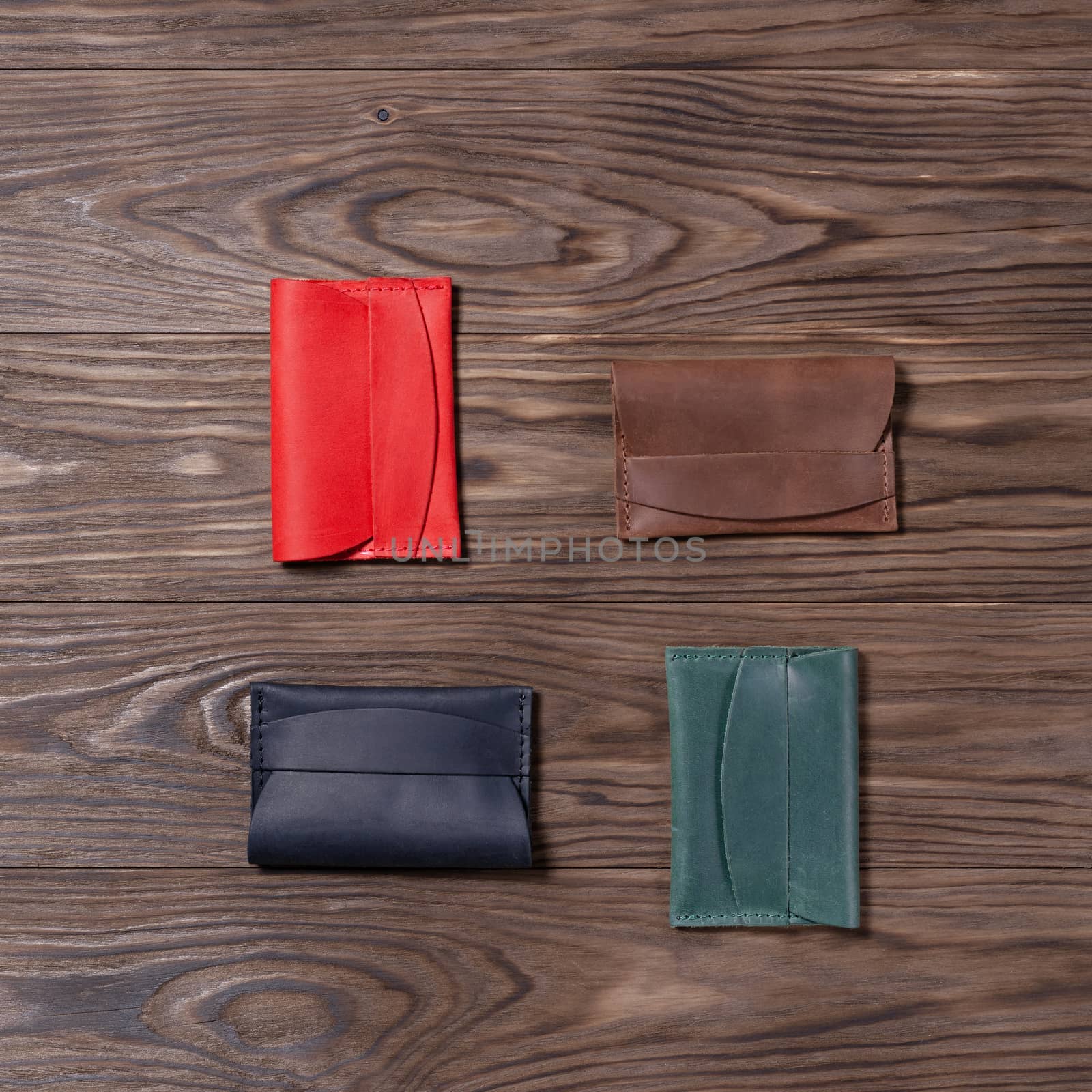 Flat lay photo of four different colour handmade leather one pocket cardholders.  Red, black, ginger and green colors. Stock photo on wooden background.