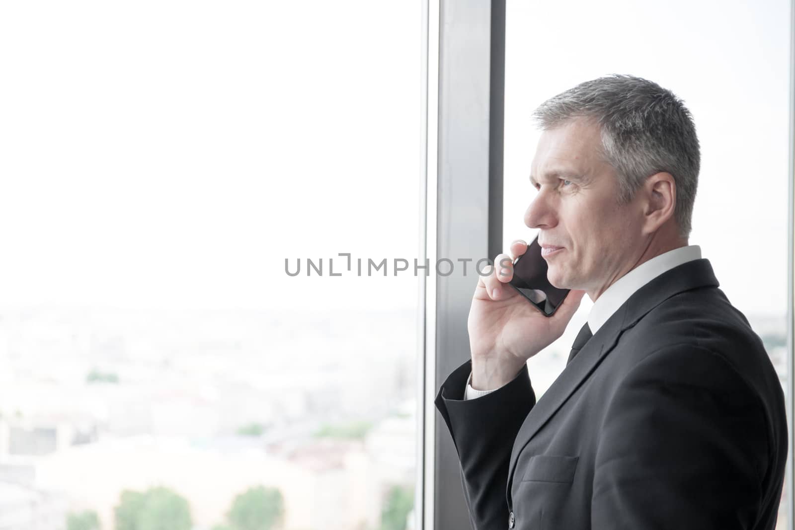 Mature businessman talking on a mobile phone standing by the window with view on city