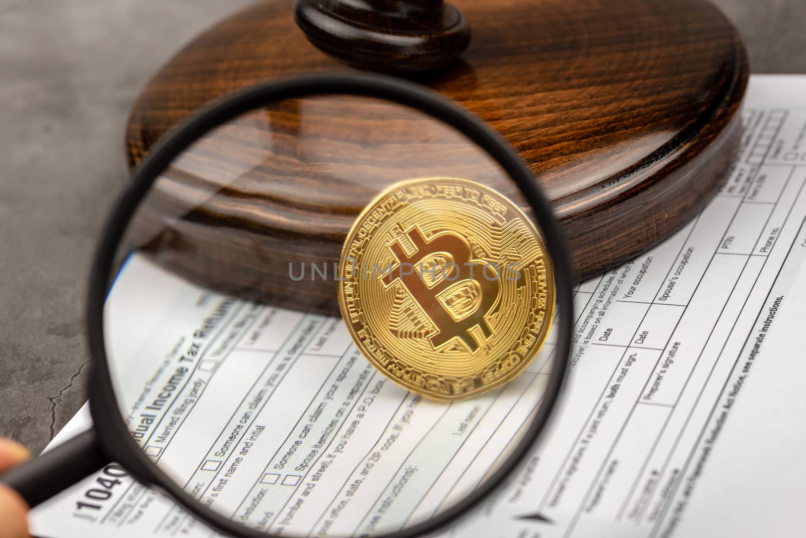 Mand hand holds magnifying glass with view of Bitcoin cryptocurrency in ront of gavel.Tax paying concept. 