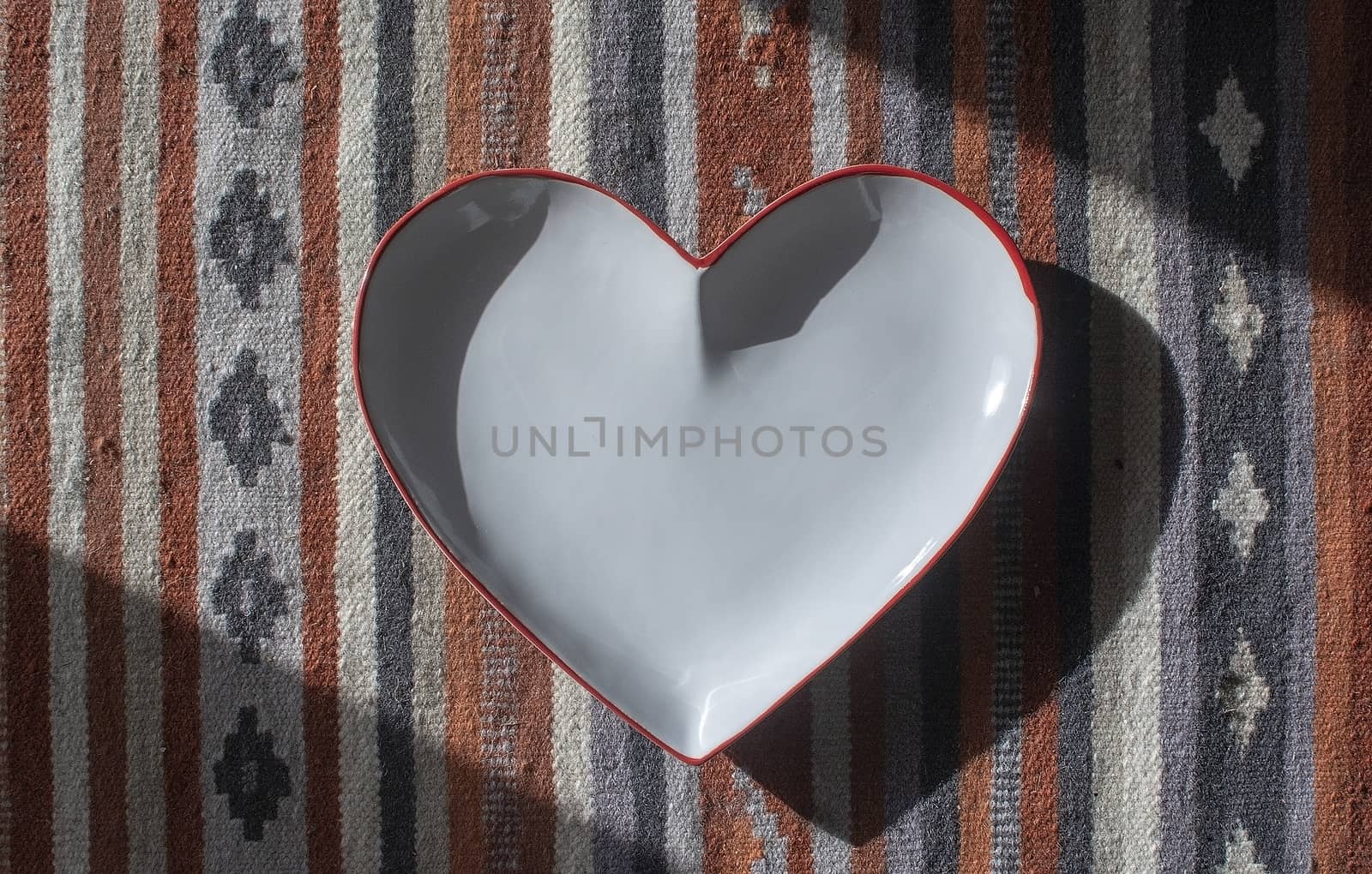 Heart shaped white plate with red edge on rustic rug with shadows decorative background.